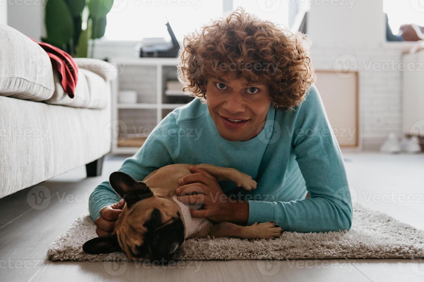 Handsome young curly man petting his cute dog and smiling while lying on the floor at home together photo