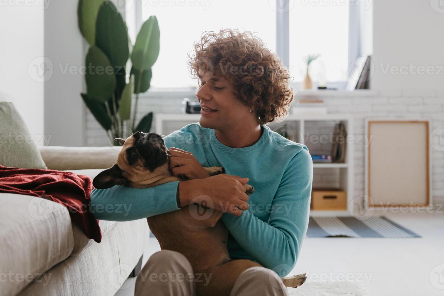 Handsome young curly man holding his cute dog and smiling while relaxing at home together photo