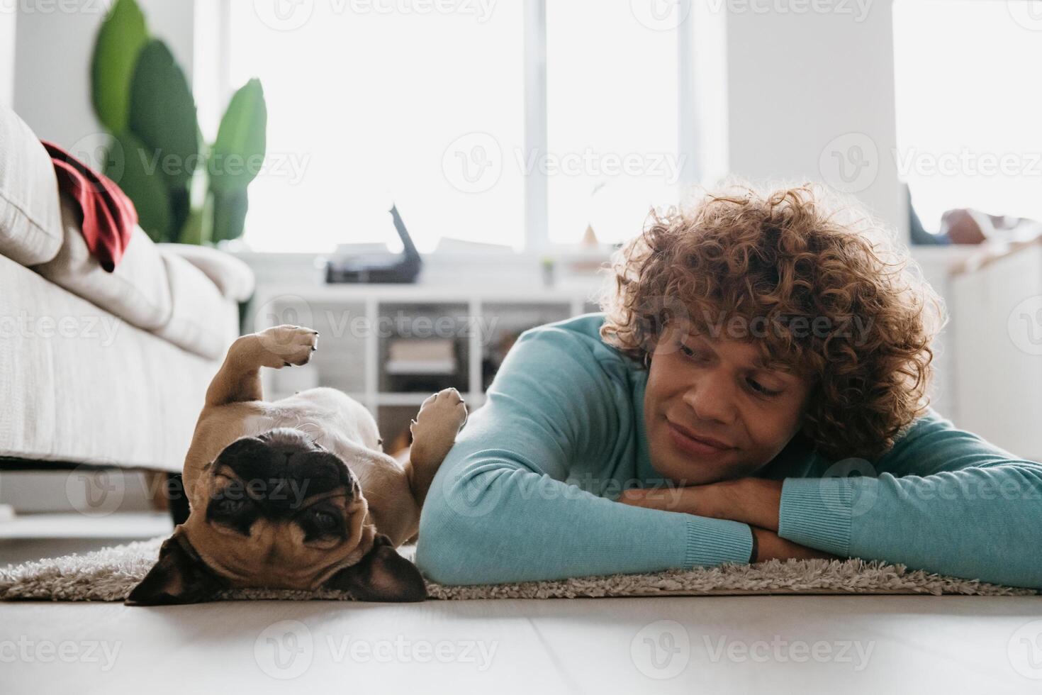 Handsome young curly man looking at his cute dog sleeping while lying on the floor at home together photo