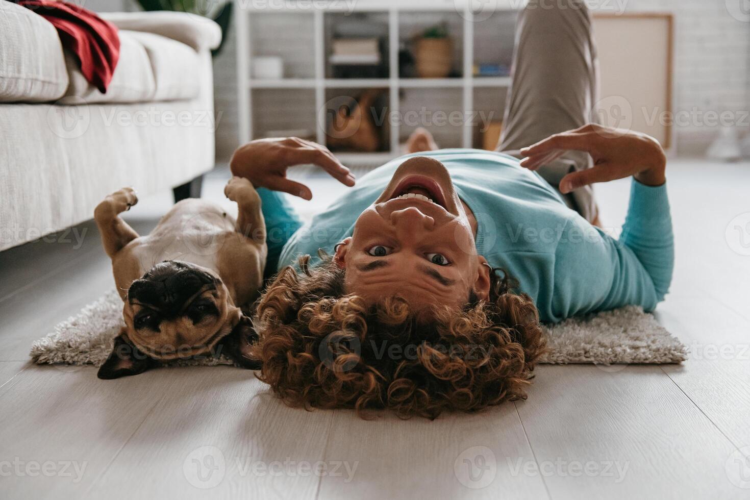 Playful young man looking at camera and smiling while lying on the floor with his cute funny dog photo
