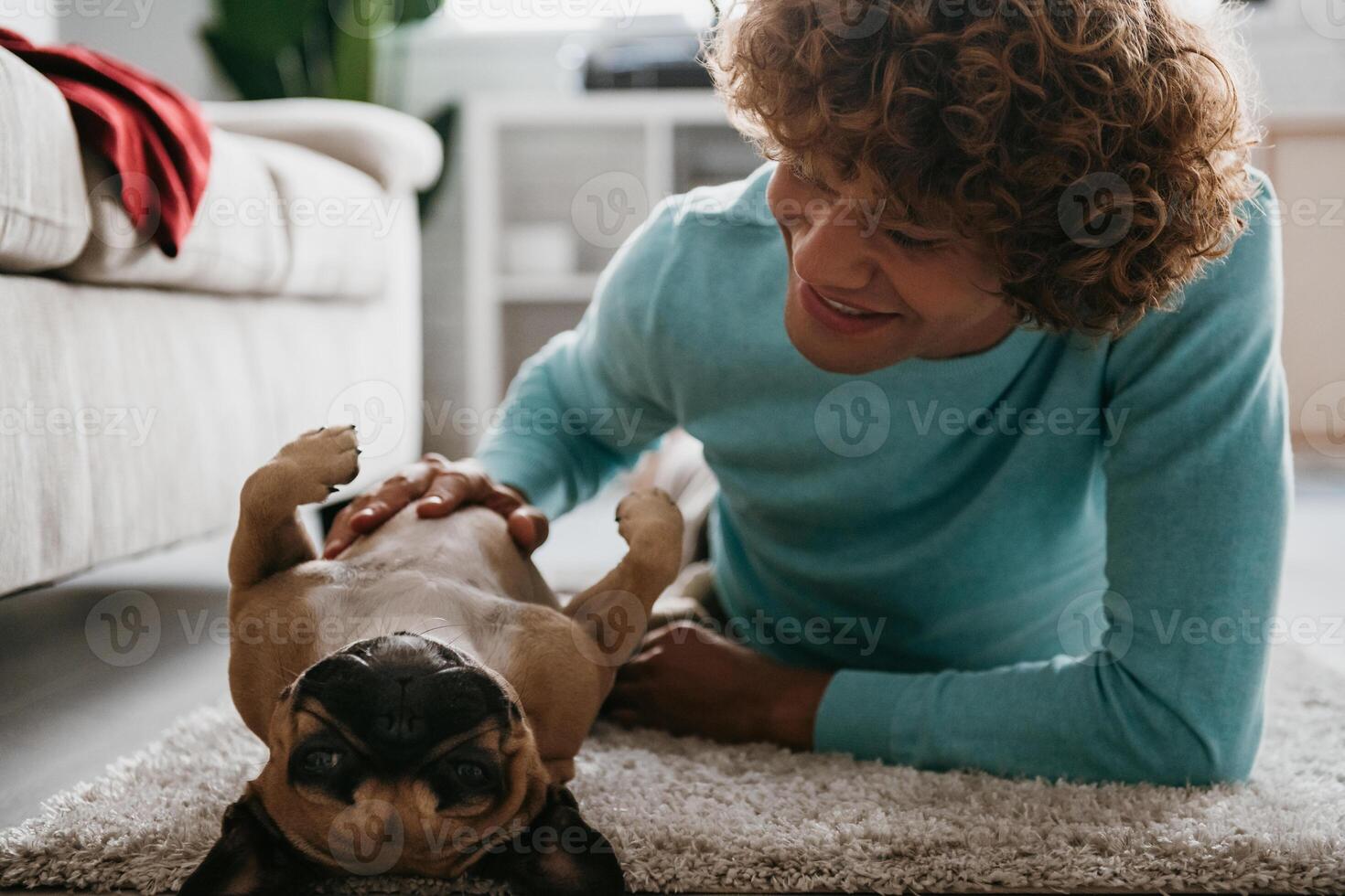 Cheerful young curly man petting his cute dog while lying on the floor at home together photo
