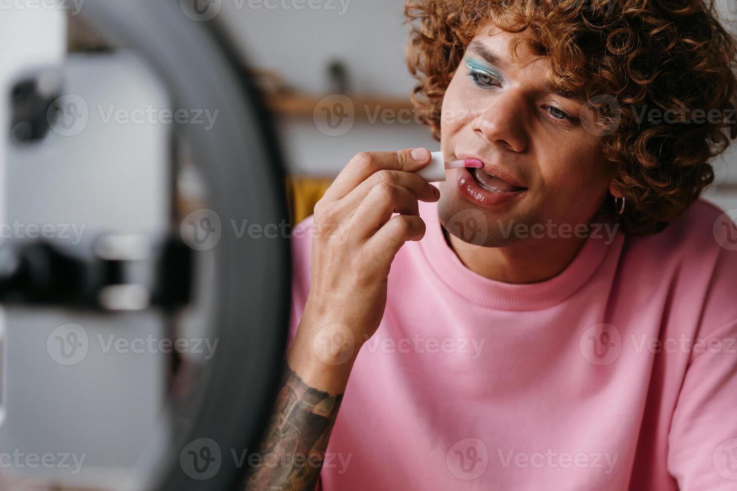 Confident young gay man applying lipstick while streaming online make-up vlog from home photo