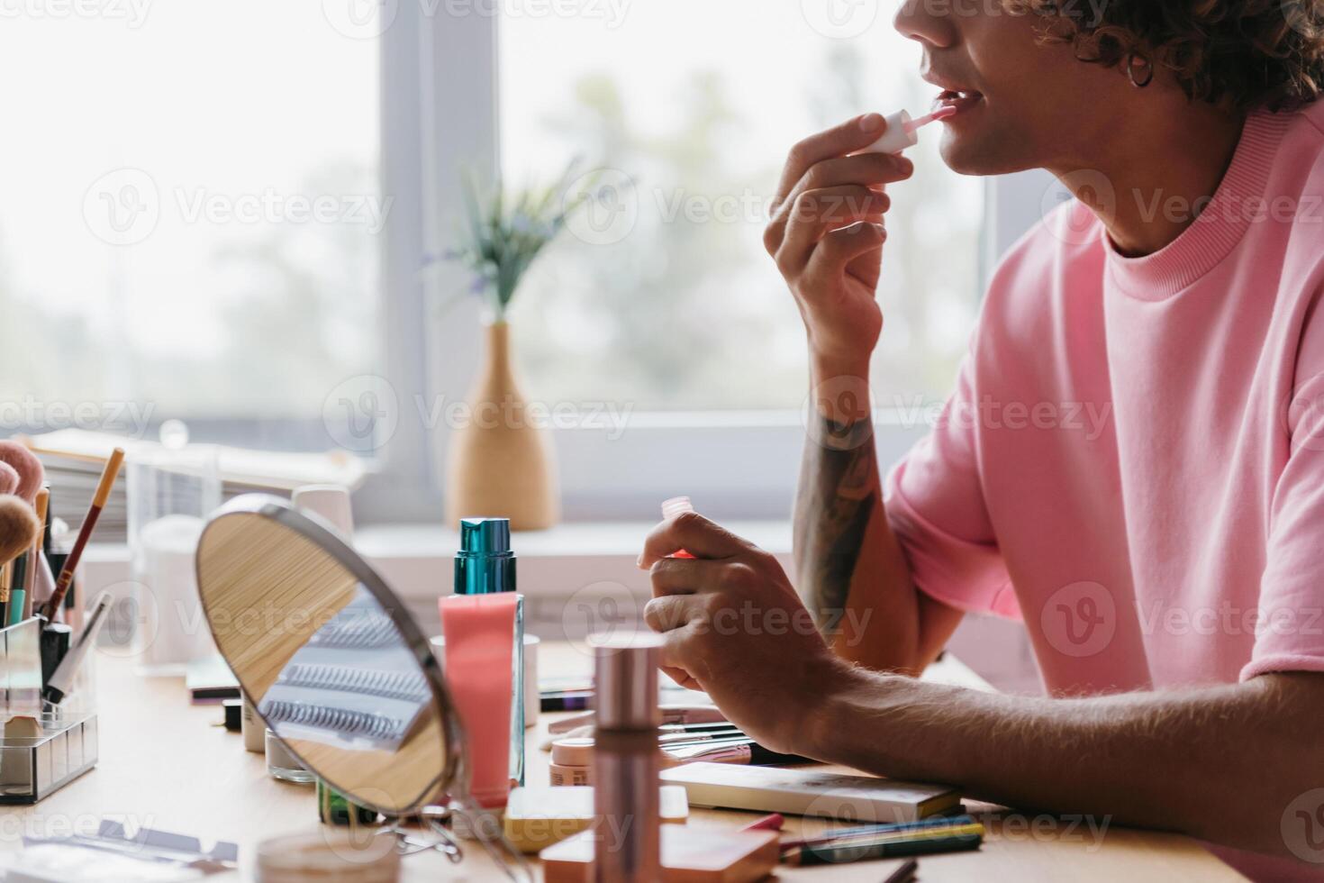 Close-up of young gay man applying make-up while sitting near the window at home photo
