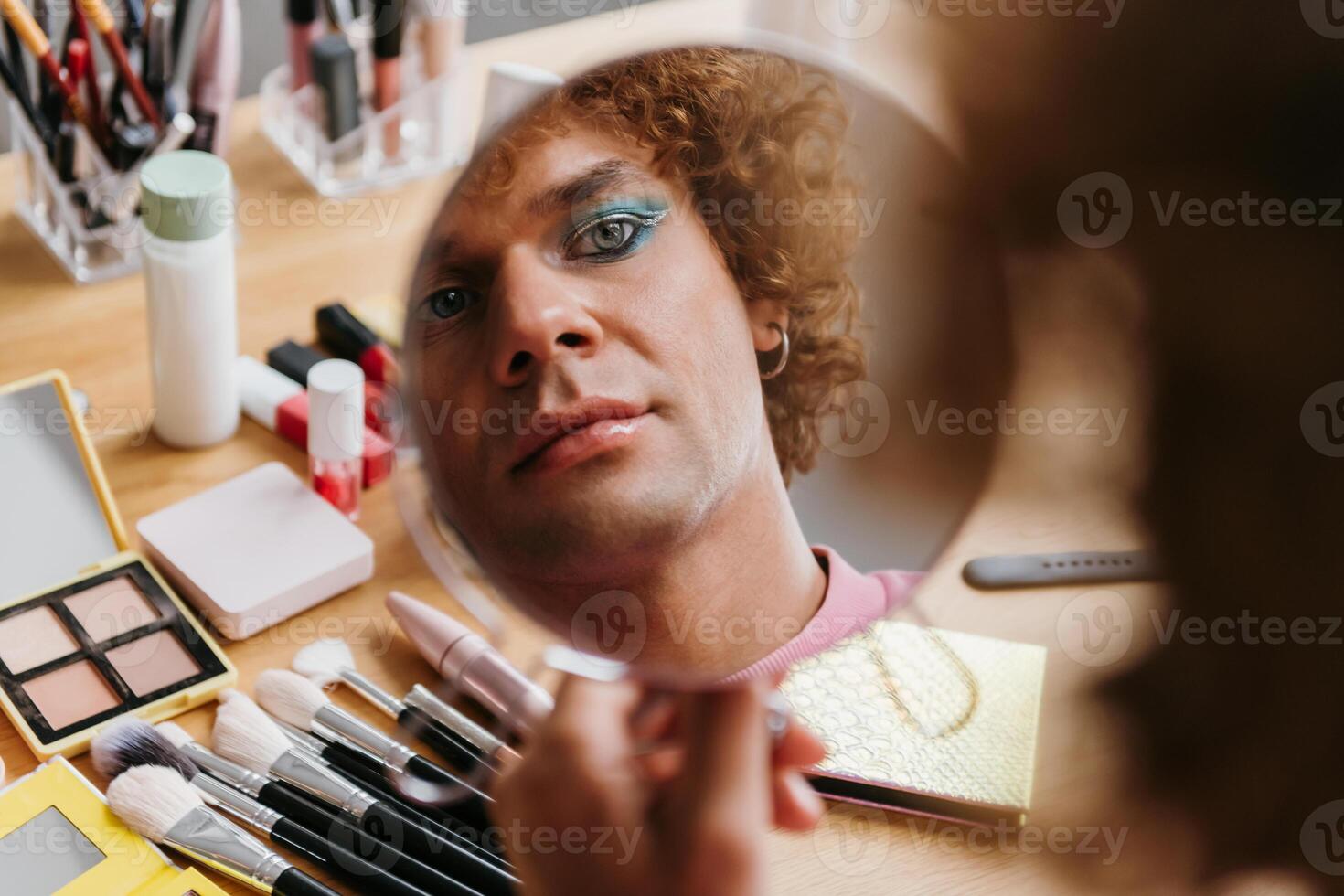 Confident young gay man looking at his reflection in the mirror while applying make-up photo