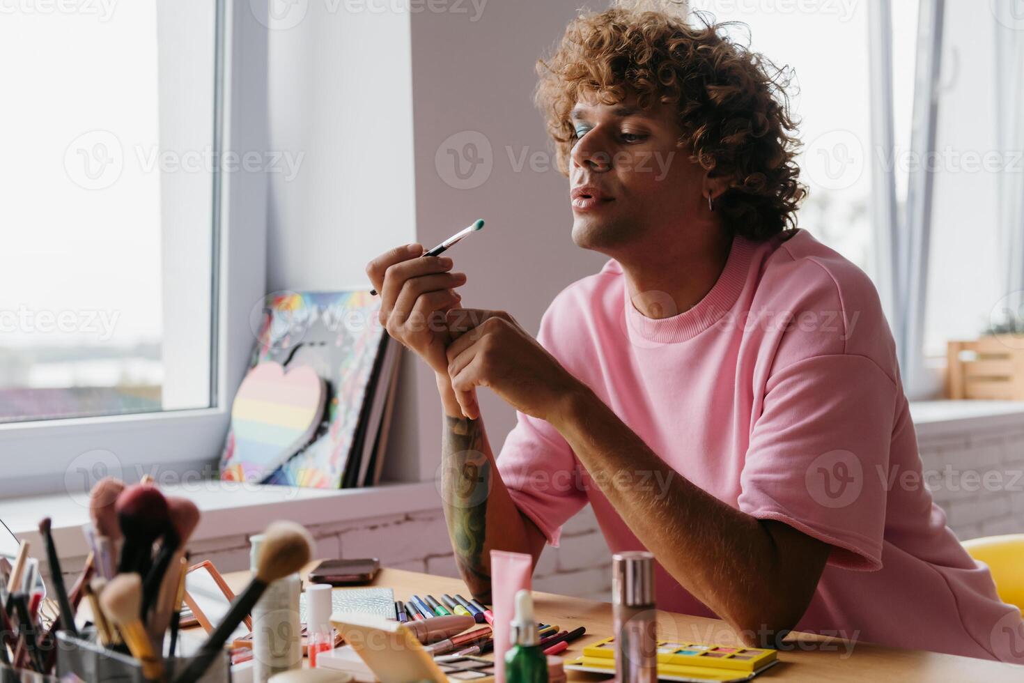 Confident young gay man blowing on eyeshadow brush while applying make-up at home photo