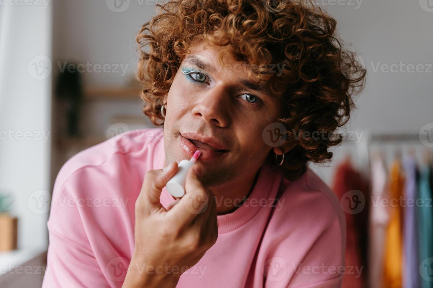 Young gay man with make-up applying lipstick and looking at camera while spending time at home photo