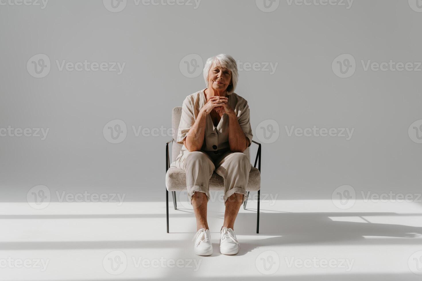 Relaxed gray hair senior woman sitting in comfortable chair and looking at camera in sunny studio photo