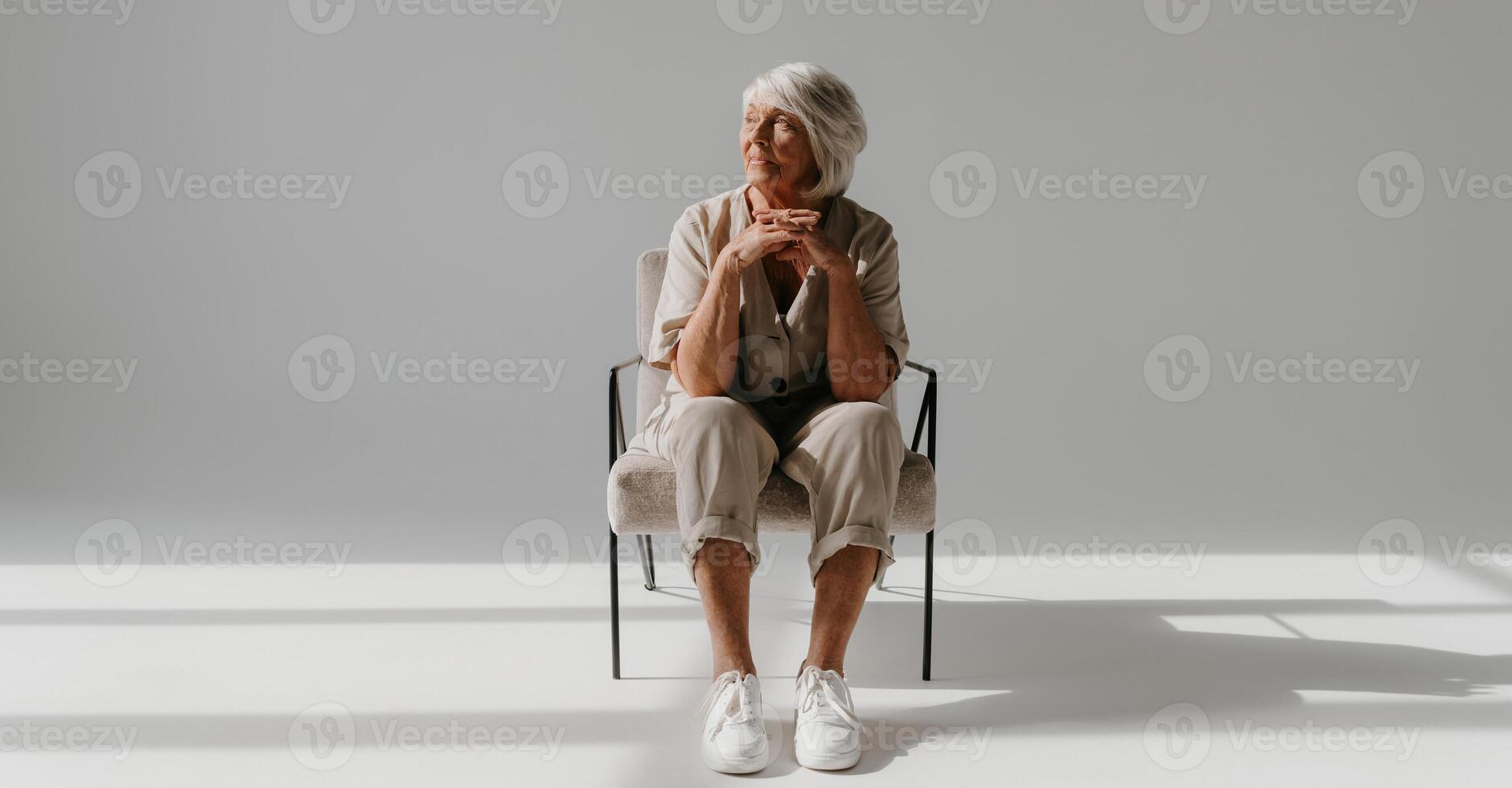Relaxed gray hair senior woman sitting in comfortable chair and looking away in sunny studio photo