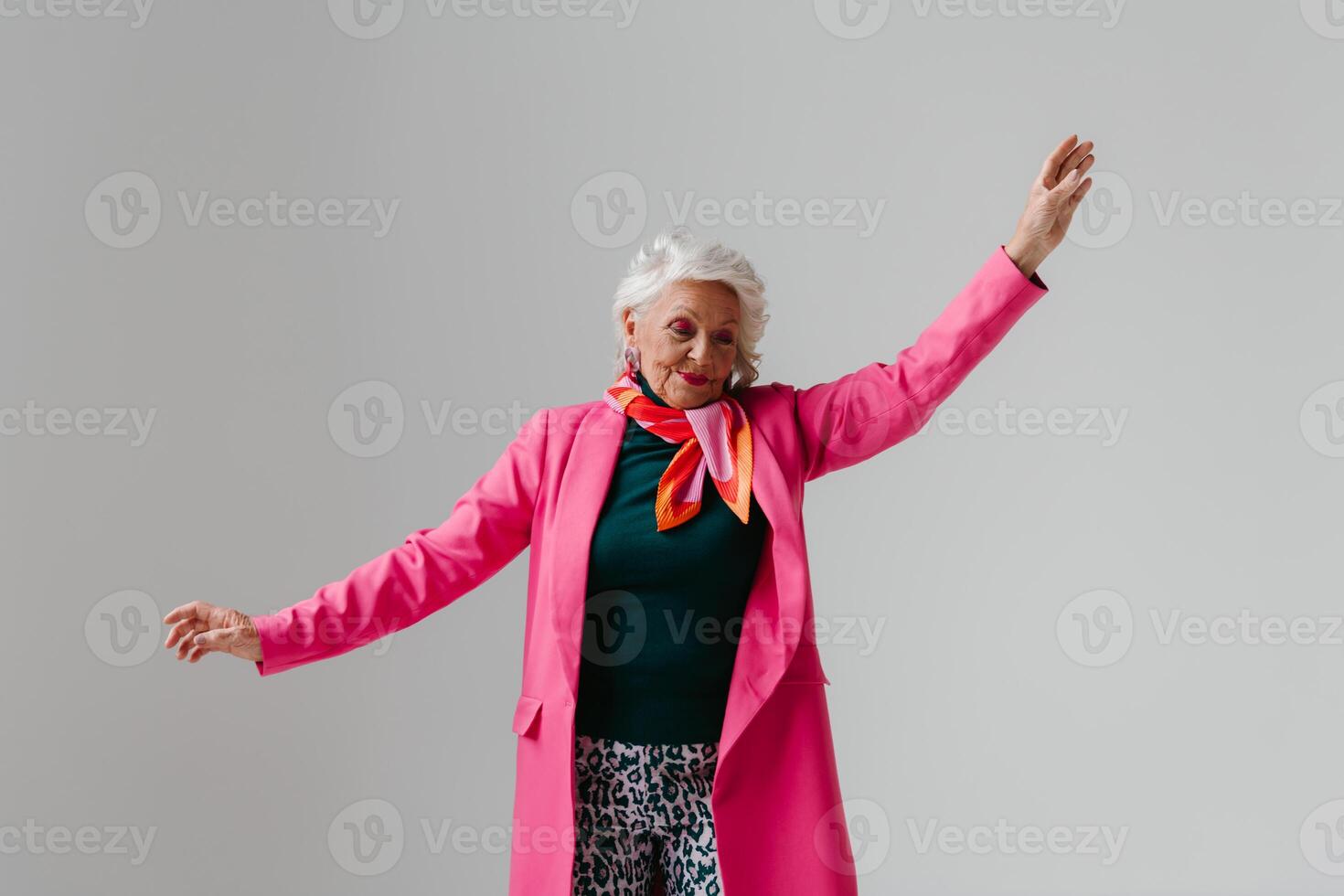 Elegant elderly woman in fashionable clothing radiating self-love while dancing on grey background photo