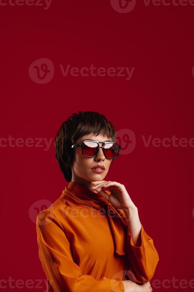 Attractive young short hair woman in trendy glasses holding hand on chin against red background photo