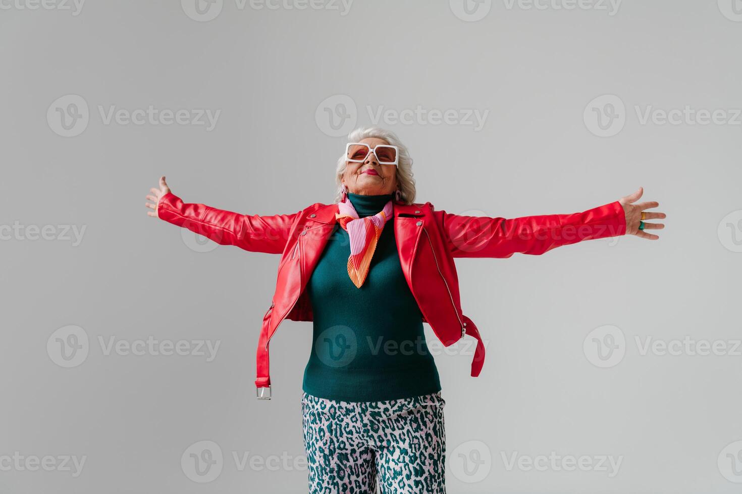 Fashionable senior woman in red leather jacket and trendy eyeglasses stretching out hands on grey background photo