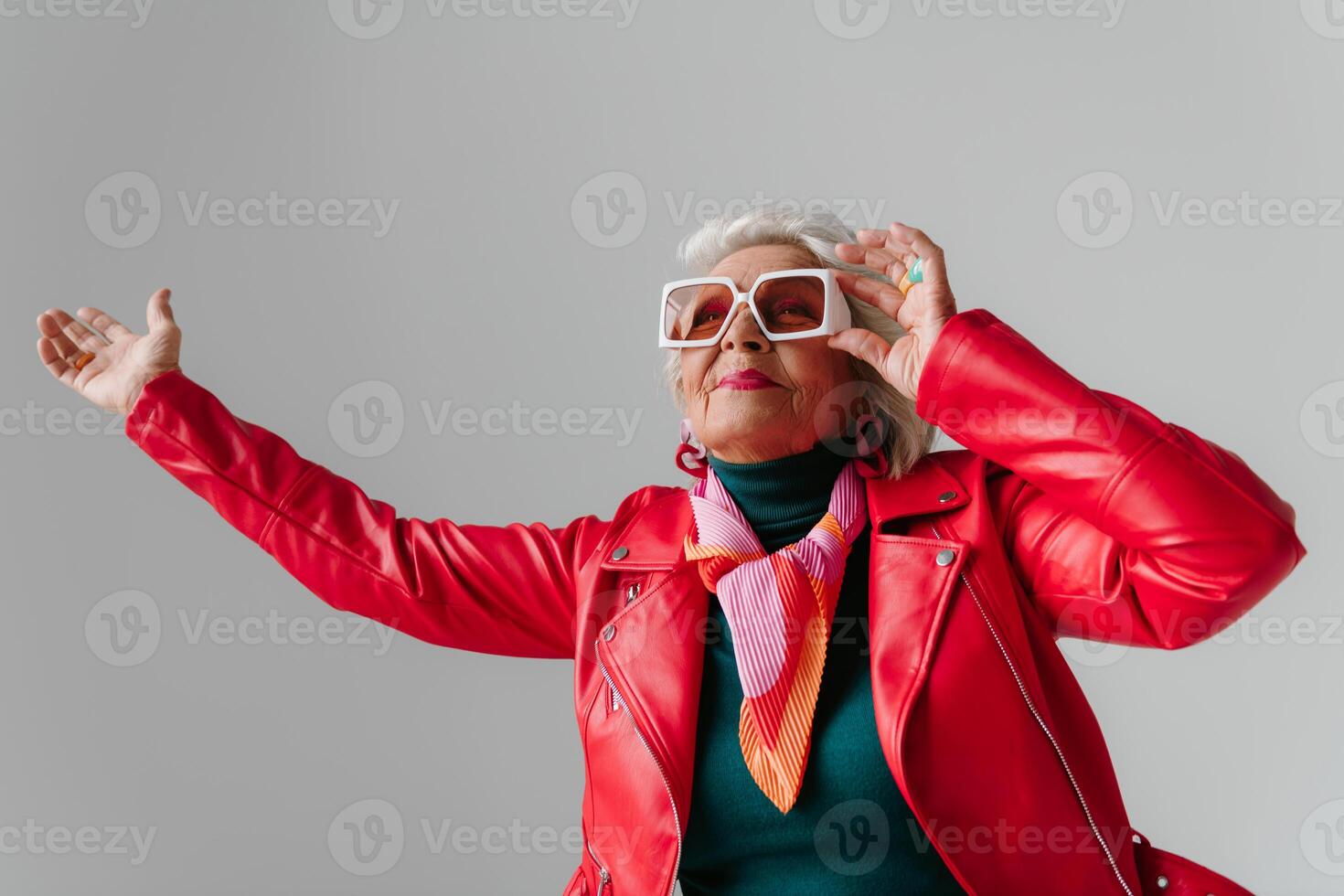 Fashionable senior woman in red leather jacket gesturing and radiating freedom on grey background photo