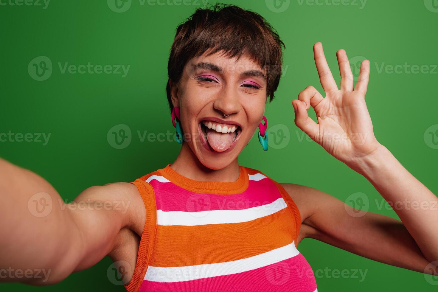 Playful young woman making selfie and gesturing while standing against green background photo