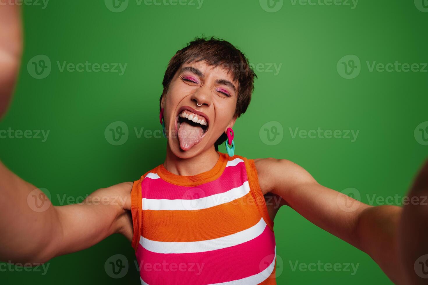 Playful young woman making selfie and sticking out tongue while standing against green background photo