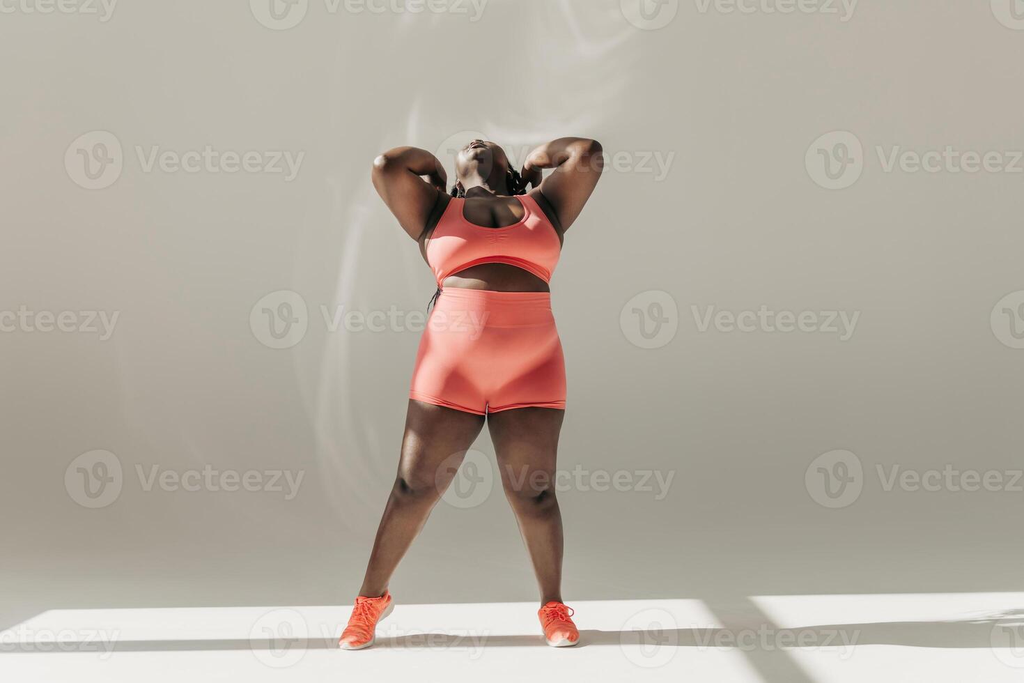 Full length of confident African plus size woman in sportswear adjusting hair while standing in fitness studio photo