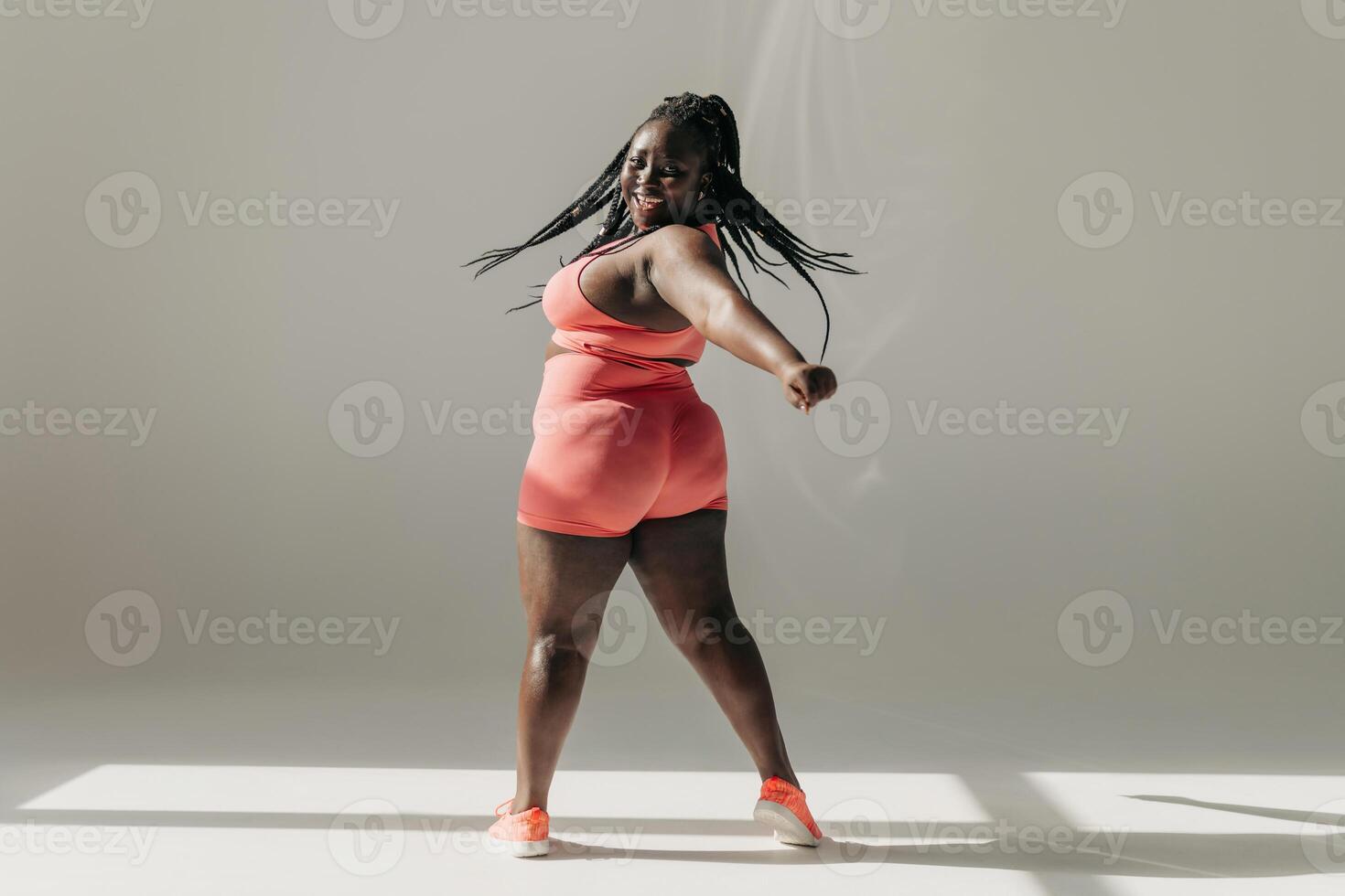 Full length of joyful African plus size woman in sportswear doing stretching exercises in fitness studio photo