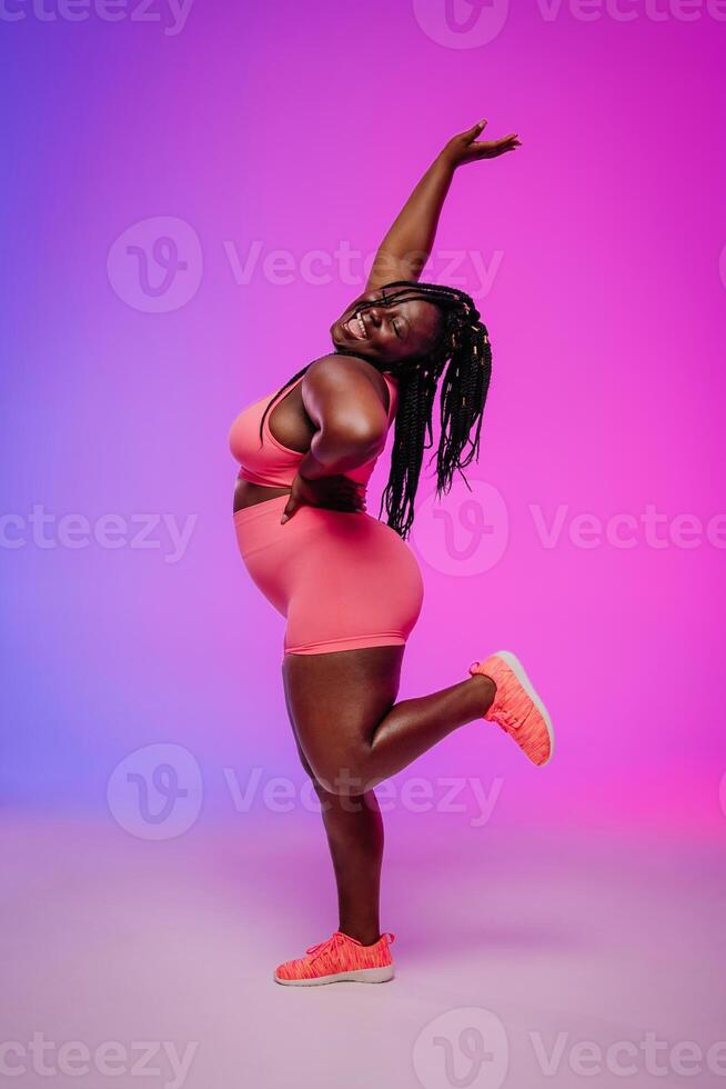 Joyful African plus size woman in sportswear dancing and radiating self-love against colorful background photo