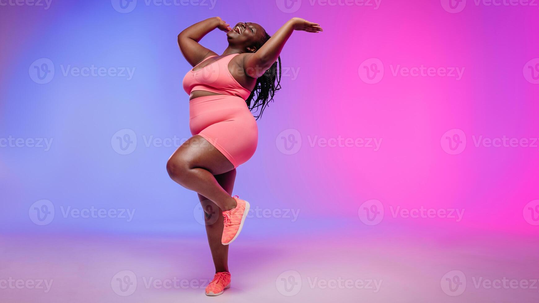 Full length of joyful voluptuous African woman in sportswear dancing and smiling on colorful background photo