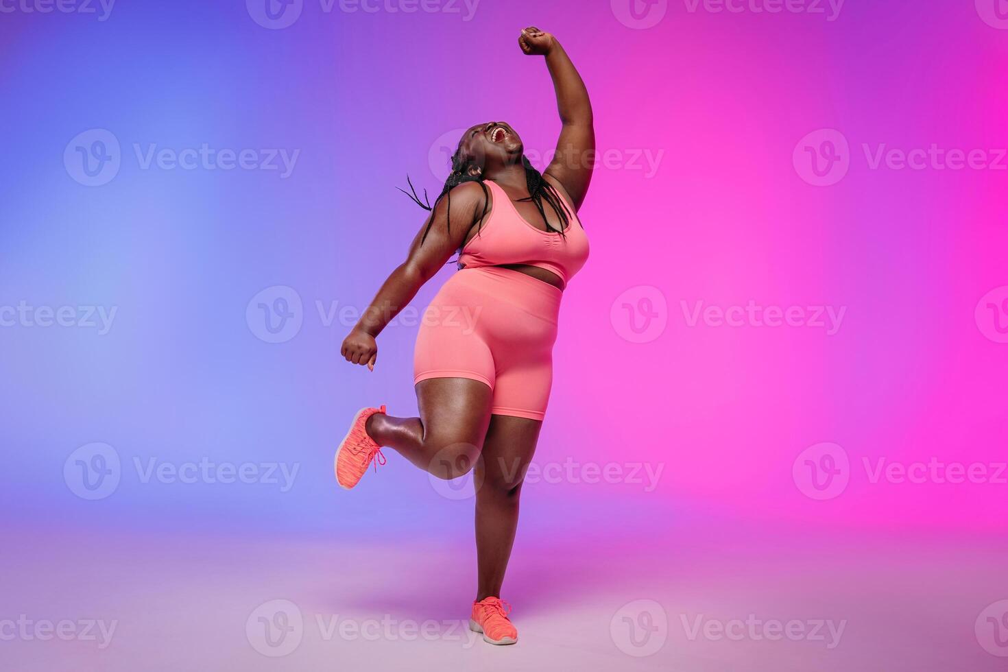 Full length of joyful curvy woman in sportswear dancing and smiling on colorful background photo