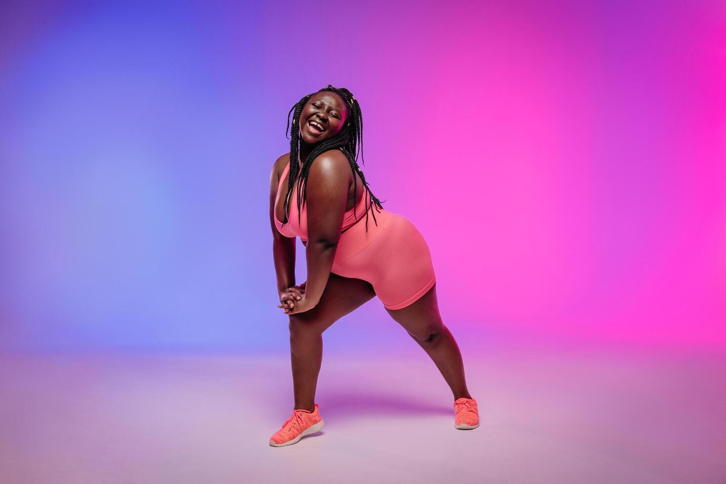 Full length of joyful plus size woman in sportswear exercising and looking confident on colorful background photo