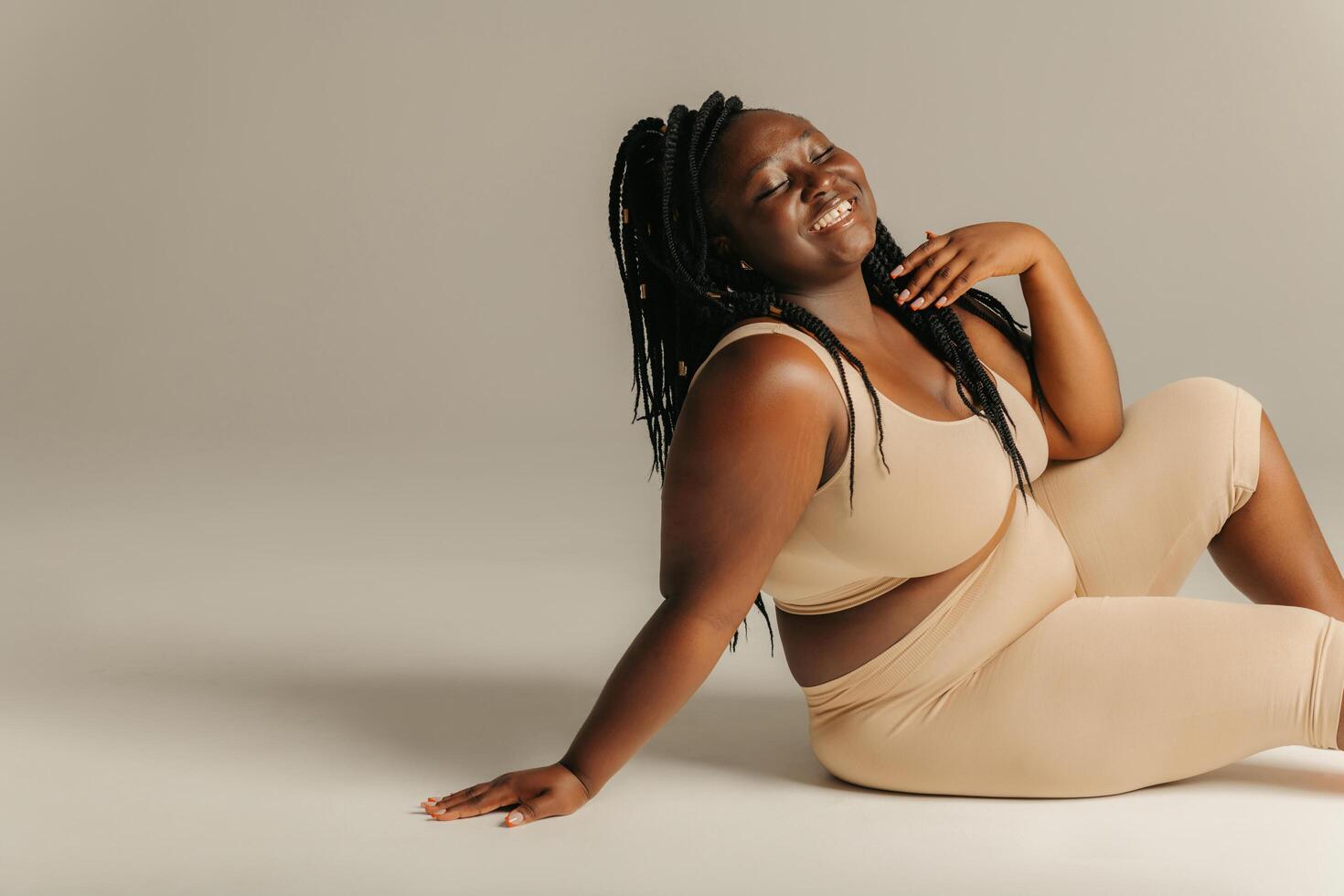 Joyful plus curvy African woman in sportswear radiating self-love while sitting against studio background photo
