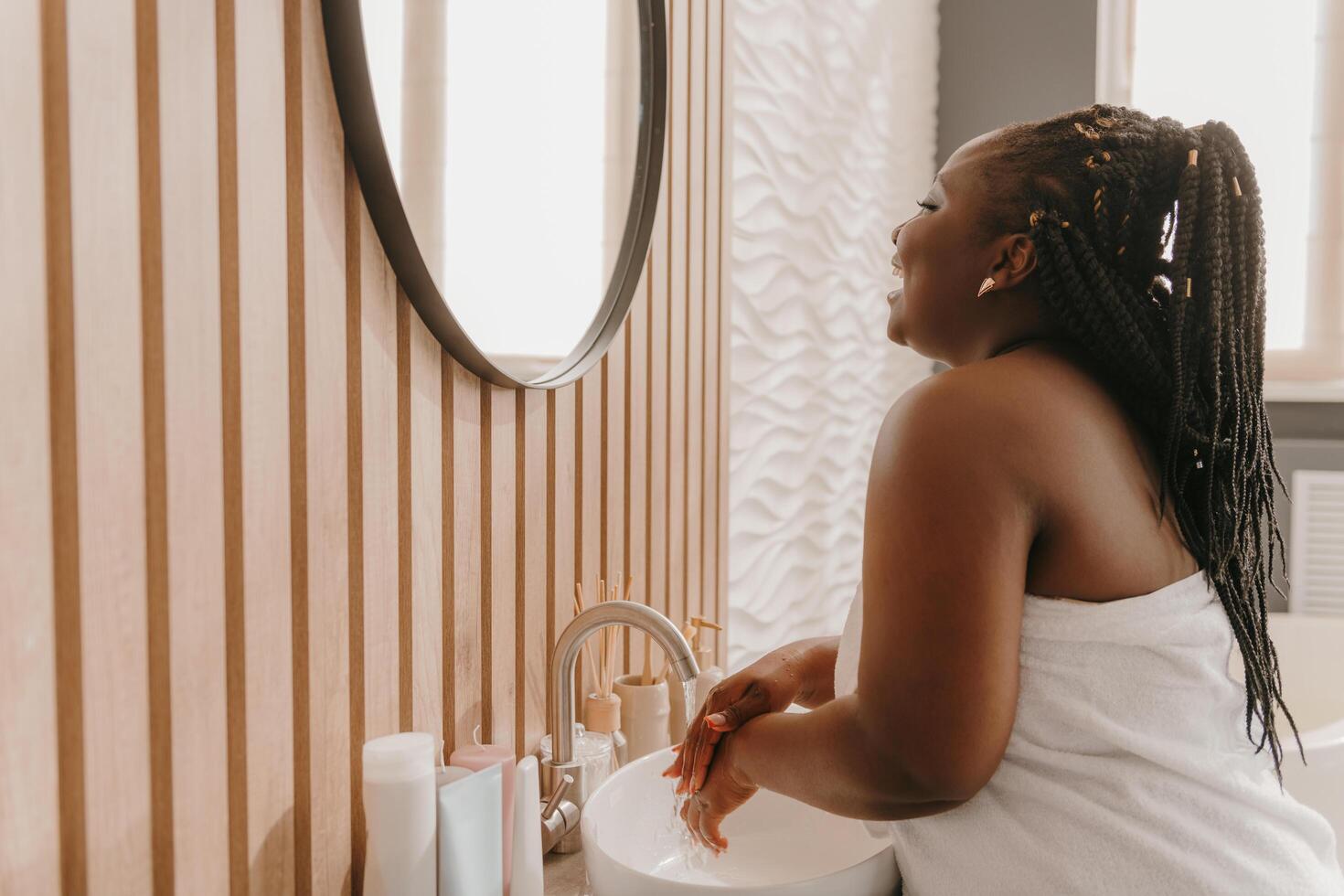 Joyful plus size African woman covered in towel washing hands and smiling at the domestic bathroom photo