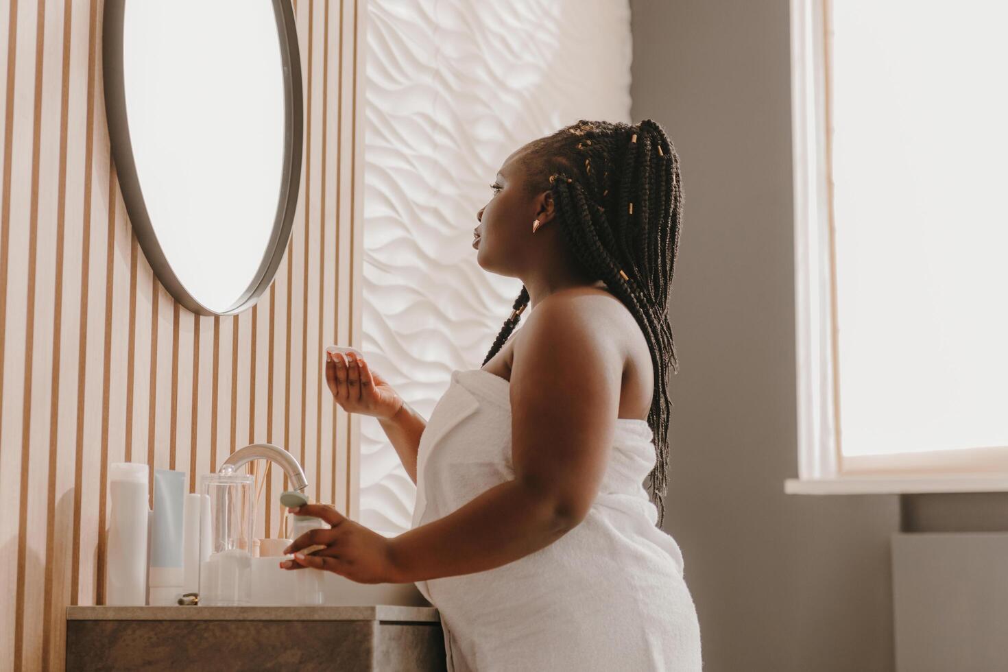 Confident curvy African woman enjoying morning routine procedures and looking at the mirror in bathroom photo