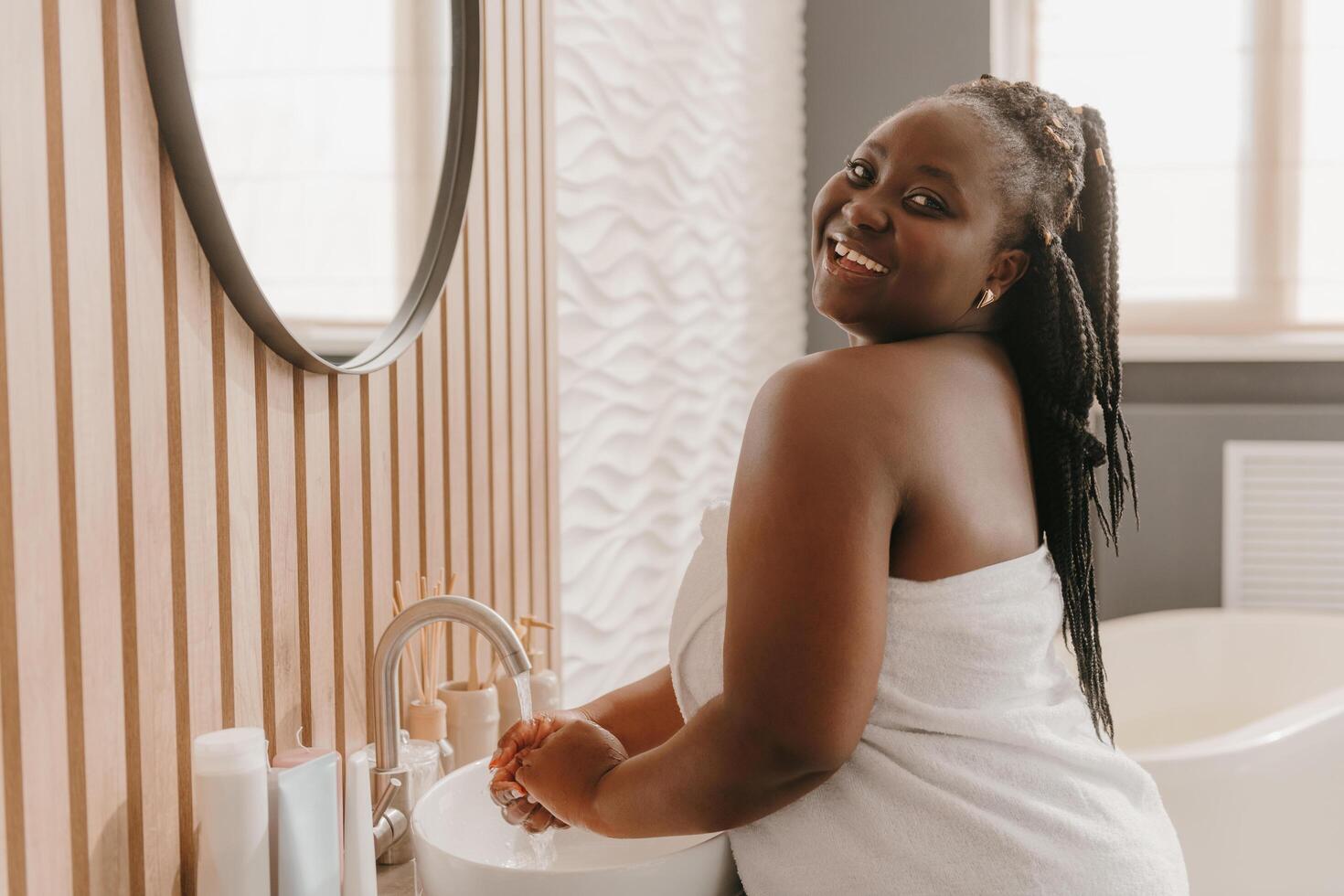 Joyful plus size African woman covered in towel washing hands and smiling at the domestic bathroom photo