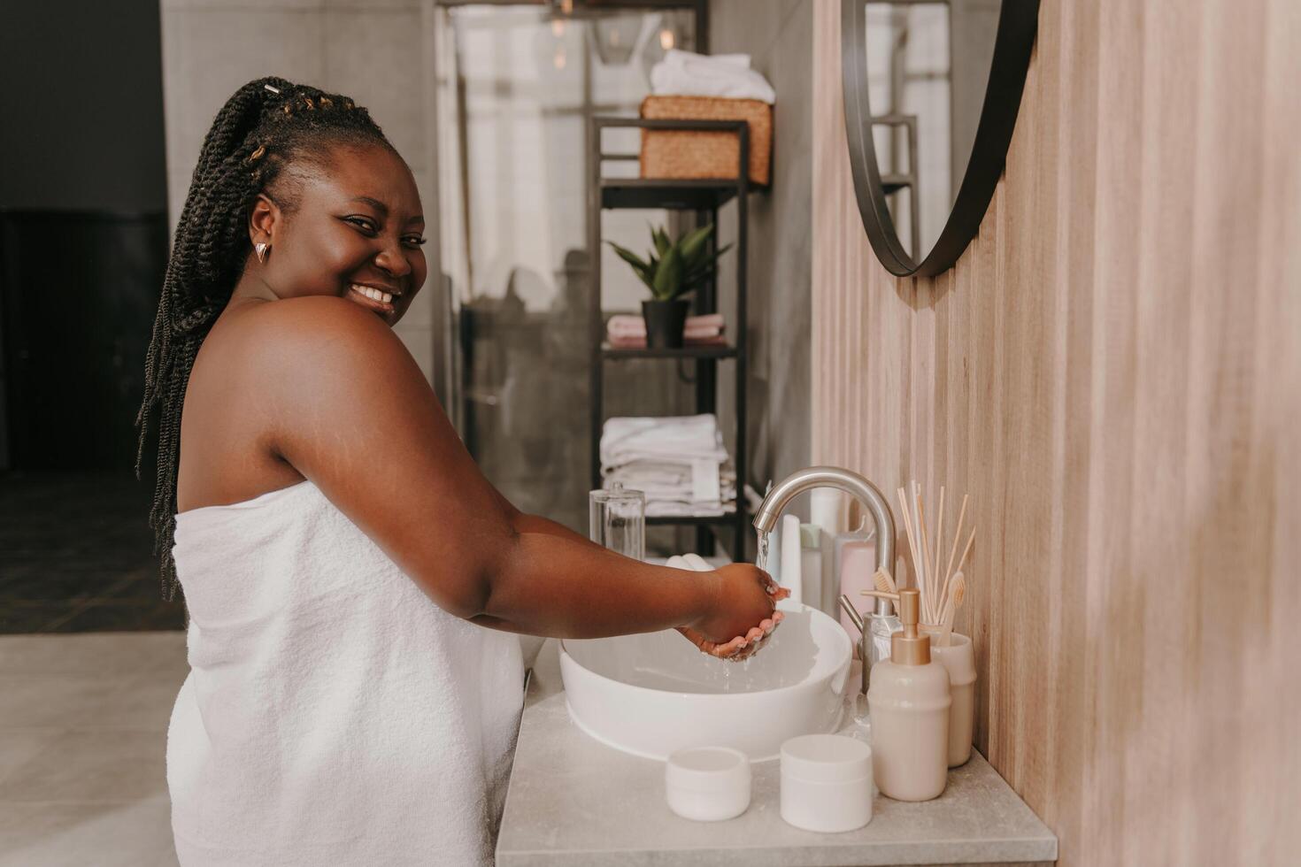 Happy curvy African woman covered in towel washing hands at the domestic bathroom photo