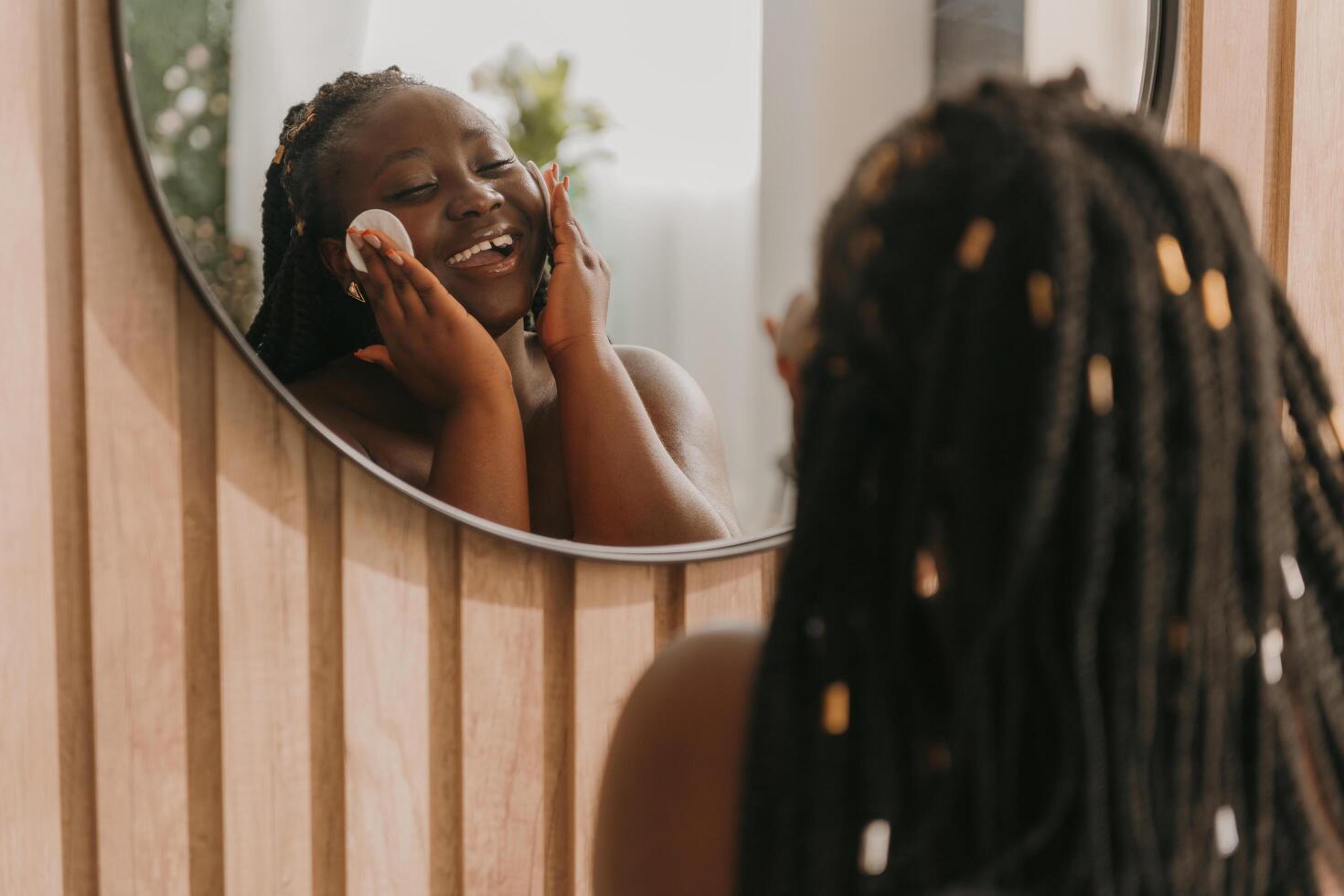 Happy plus size African woman cleaning face with cotton pad and looking at the mirror in bathroom photo