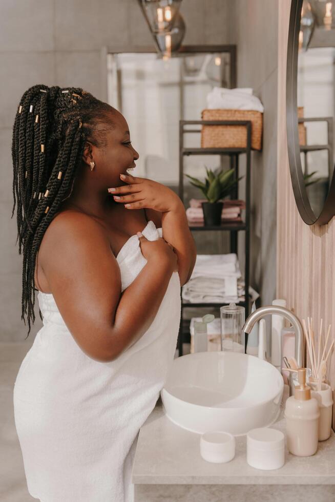 Confident plus size African woman touching face and radiating self-love while looking at the mirror in bathroom photo