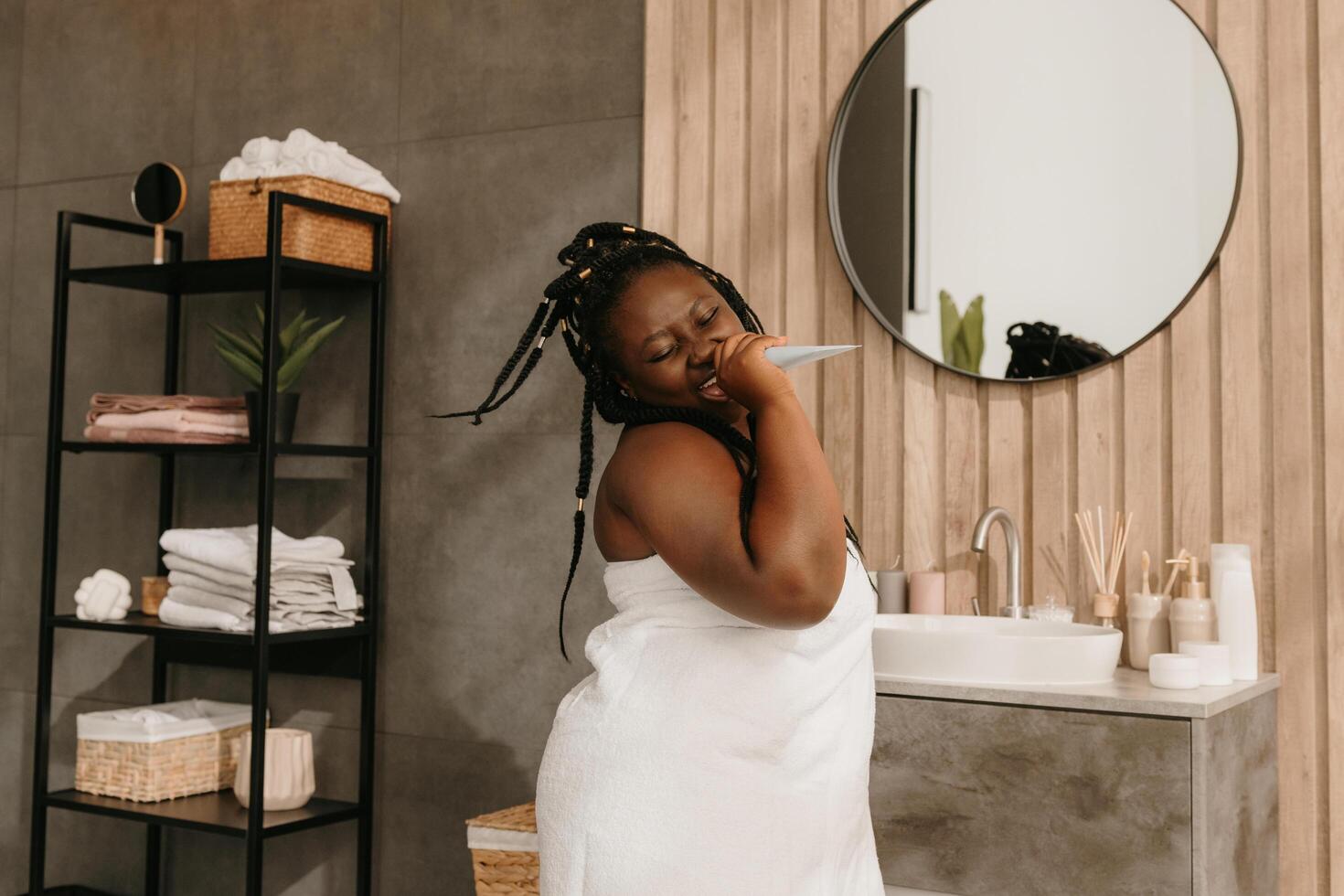 Joyful curvy African woman holding cosmetic bottle like microphone while singing in bathroom photo