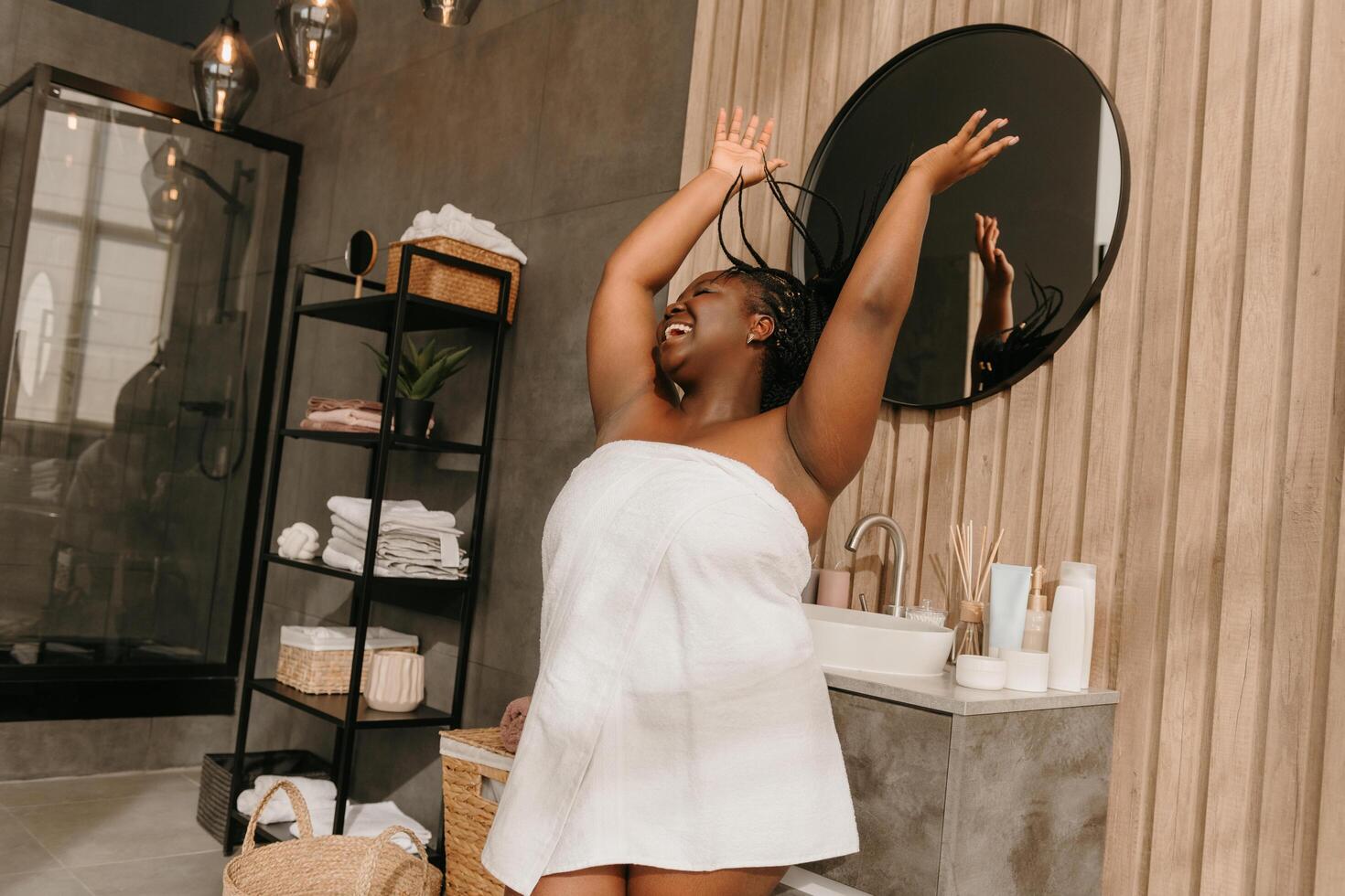 Joyful curvy African woman covered in towel playing with her braided hair and smiling in bathroom photo