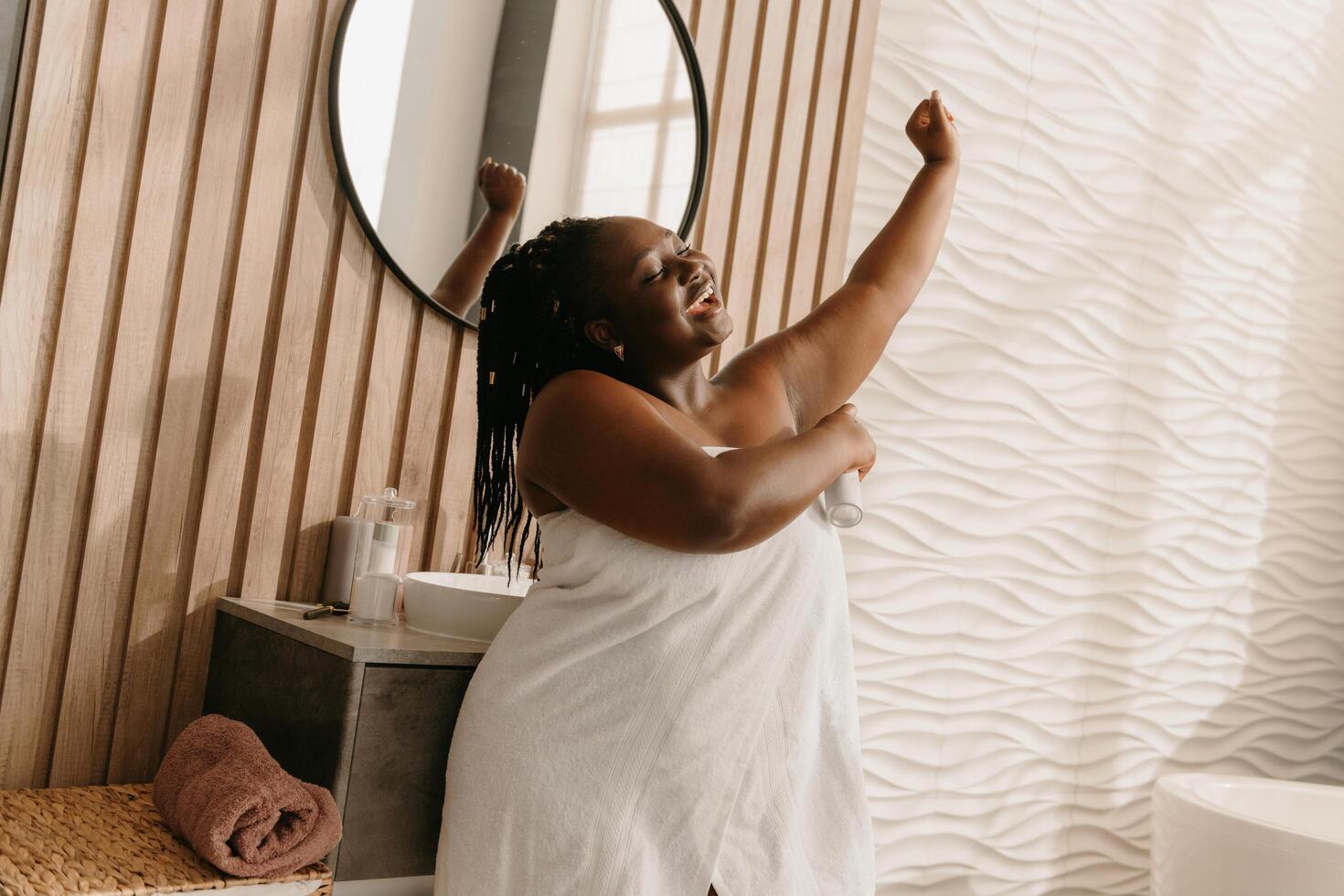 Happy plus size African woman covered in towel looking fresh and clean while spraying deodorant in bathroom photo