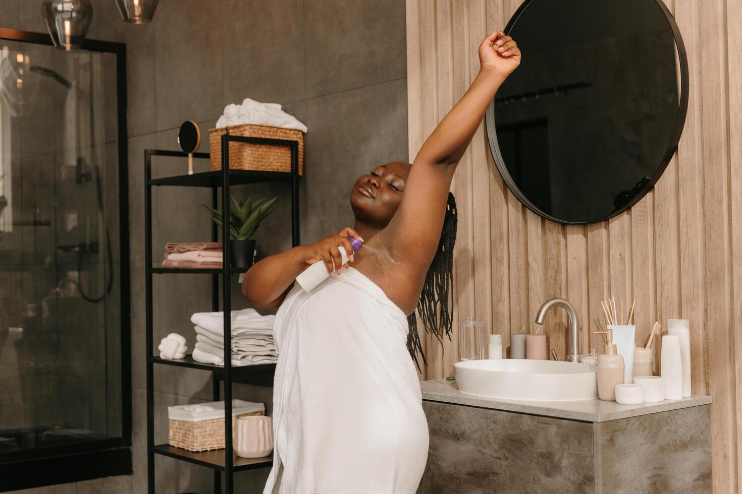 Confident plus size African woman covered in towel looking fresh and clean while spraying deodorant in bathroom photo