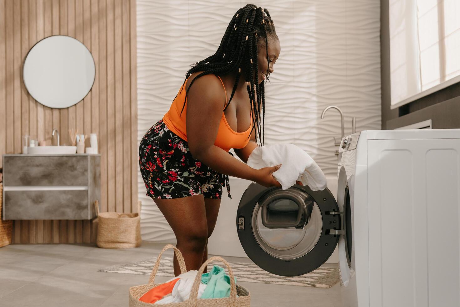 Beautiful plus size African woman loading clothes into washing machine while standing in domestic bathroom photo