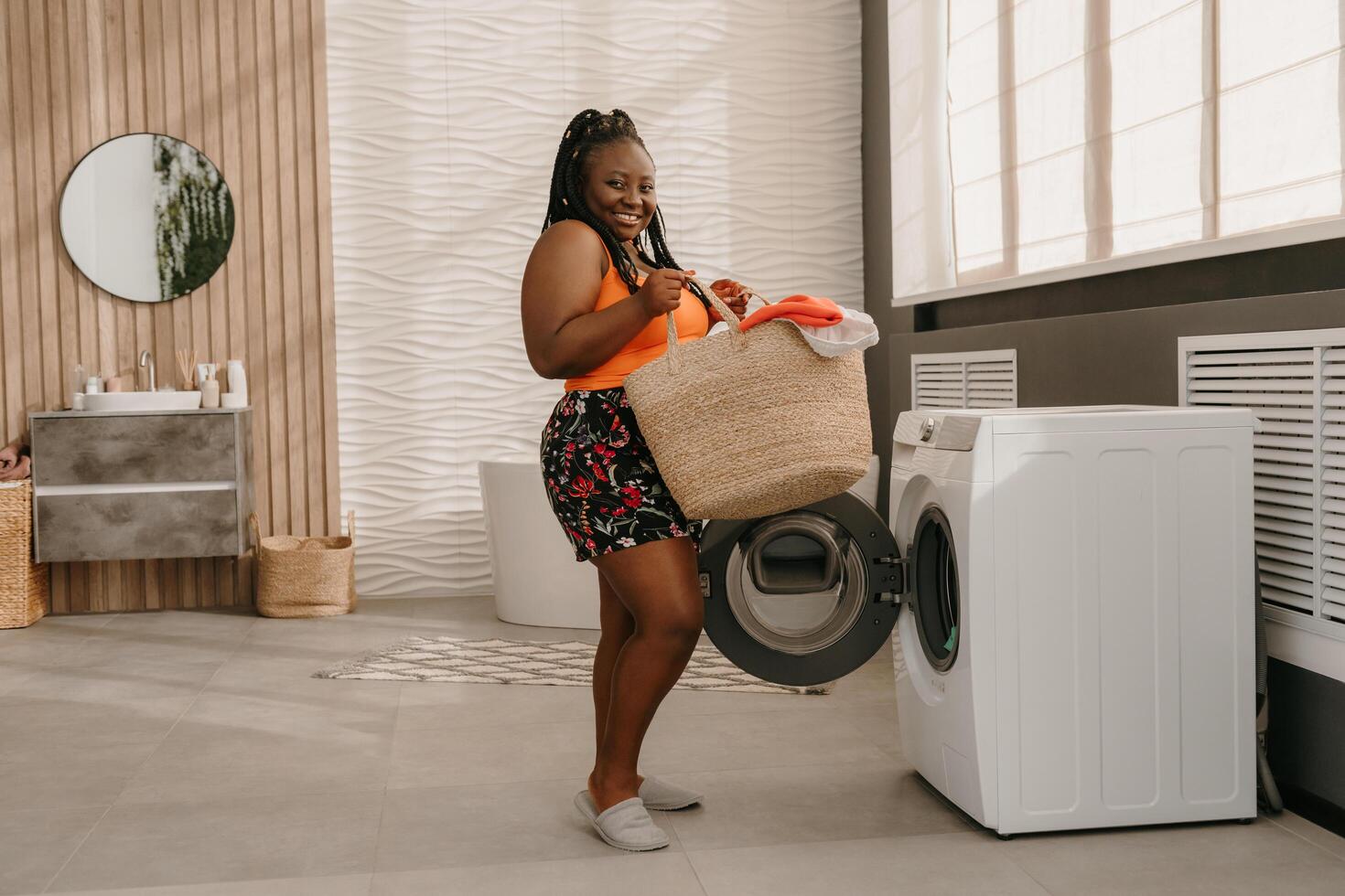 Happy plus size African woman carrying basket with clothes while standing near the washing machine in bathroom photo
