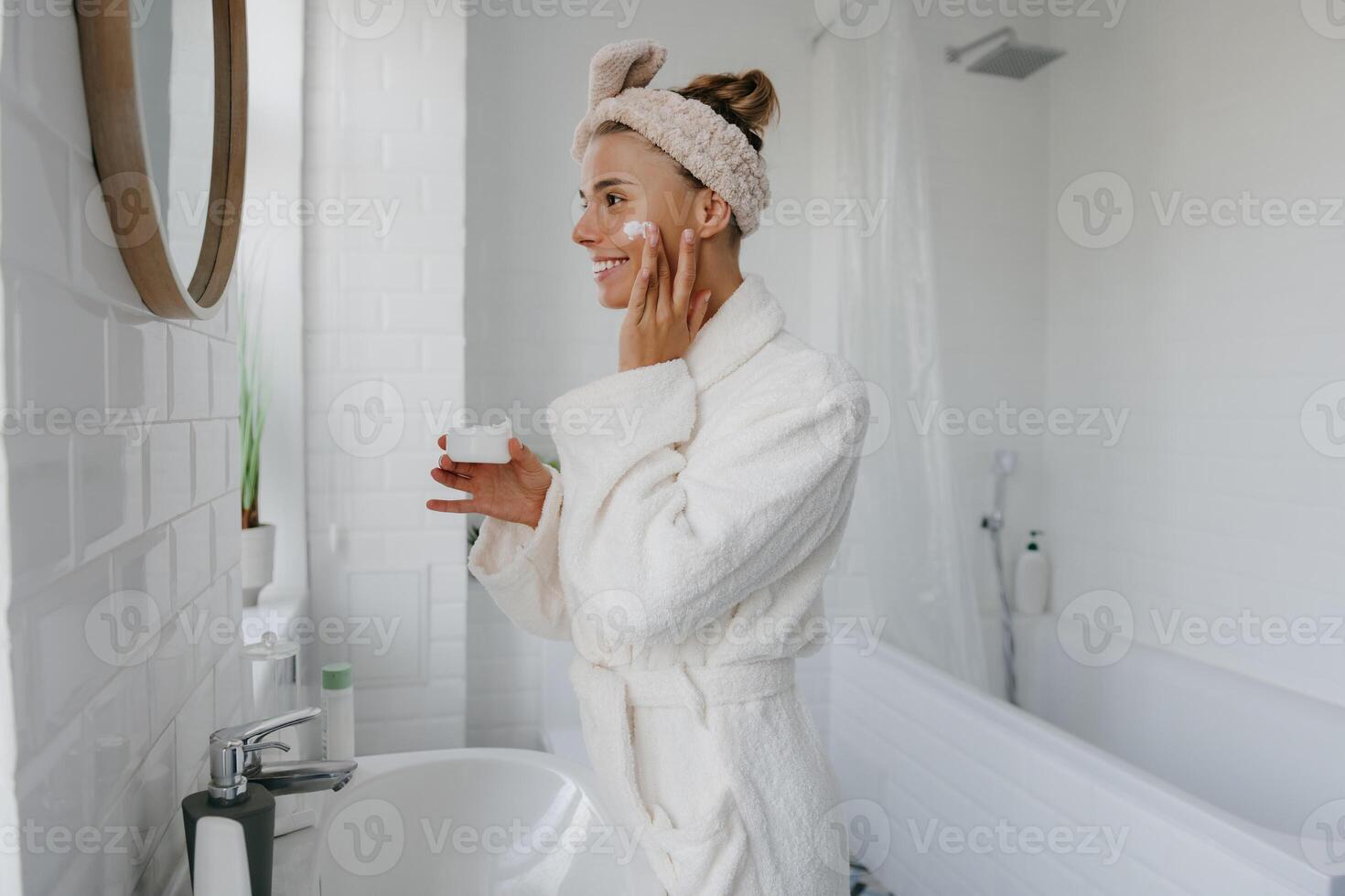 Beautiful young woman in bathrobe applying face cream and smiling while standing in bathroom photo