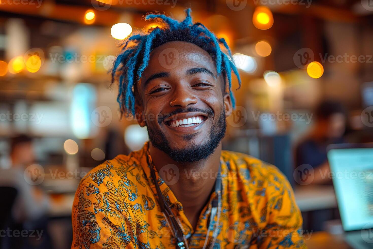 A man with blue hair and a yellow shirt is smiling. He is surrounded by people and there are two laptops in the background photo