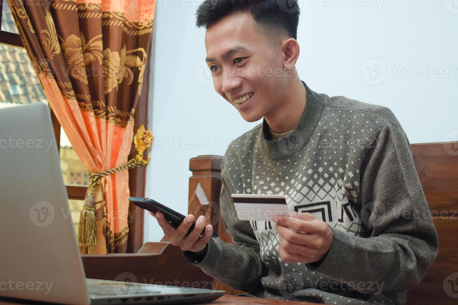 young man shopping online in e commerce, holding credit card and smartphone with laptop om table in livingroom photo