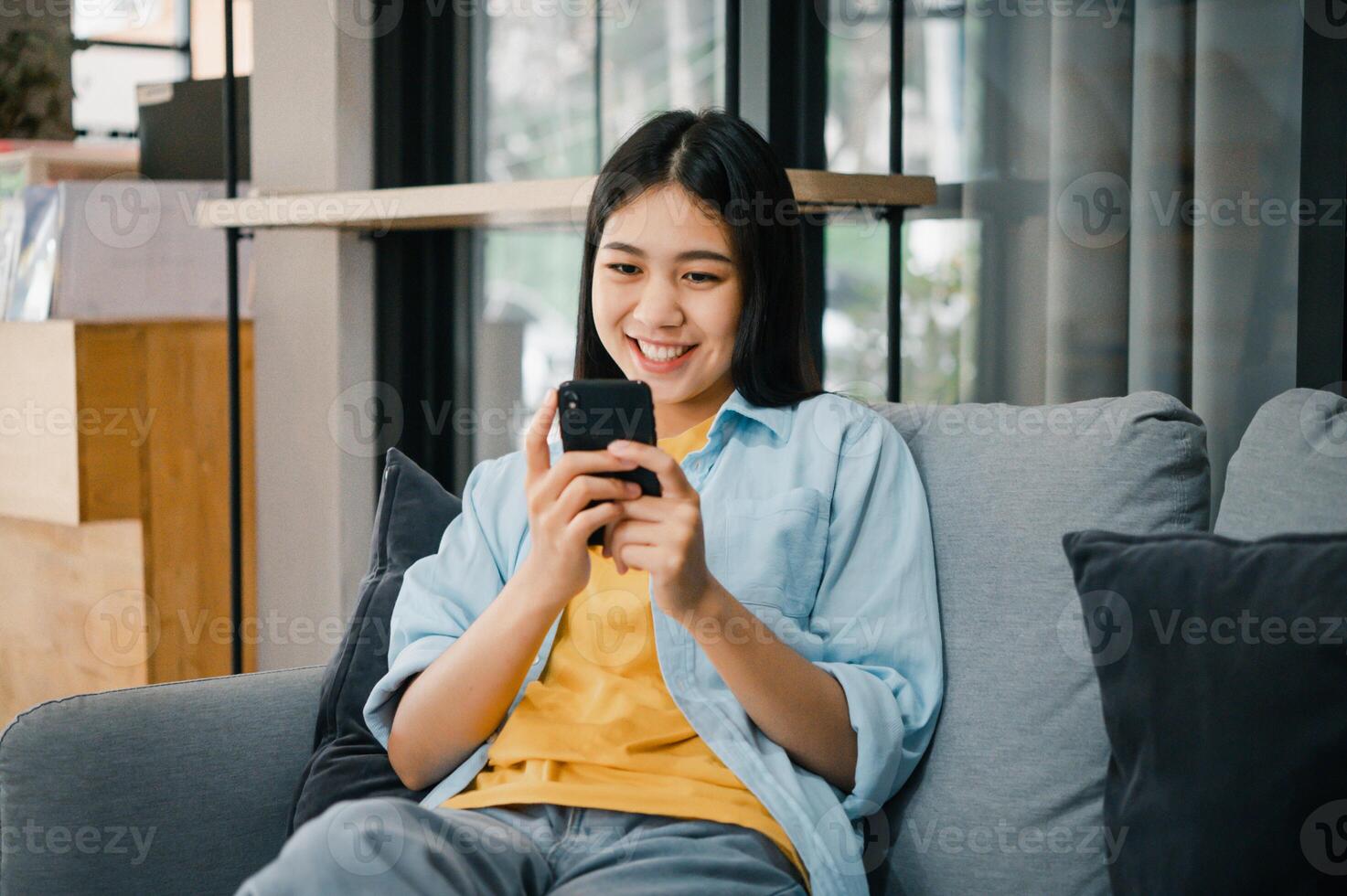 Smiling young asian woman using mobile phone checking social media,app playing game, shopping online, ordering delivery relax while sitting on a couch at home with laptop computer photo