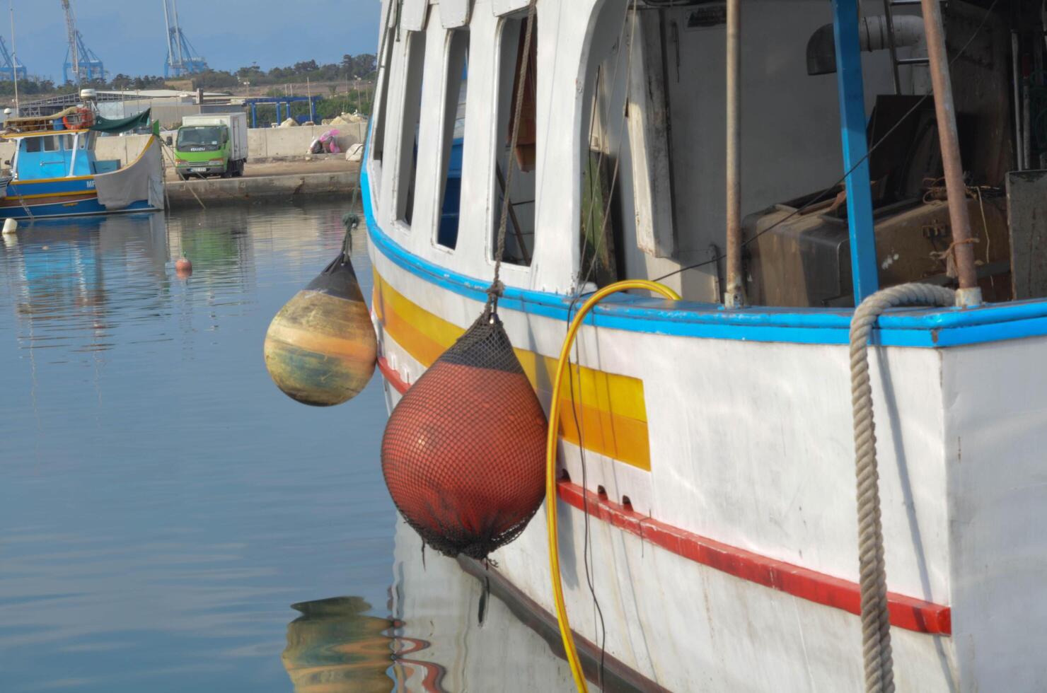 A close-up view of two orange buoys securely fastened to the side of a small fishing boat, providing protection while docked. The vibrant buoys contrast with the boat's hull, showcasing the practical photo