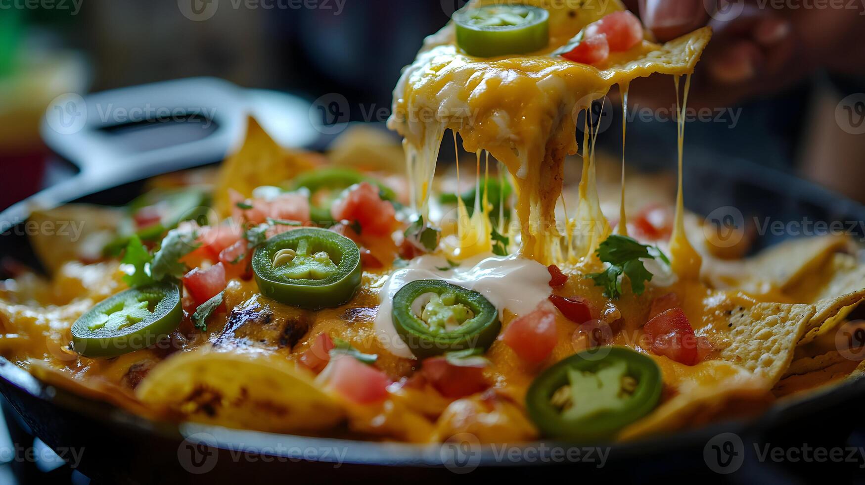 A hot skillet of nachos with cheese stretching between chips as someone lifts one out, the melted cheese glistening, with toppings like jalapenos, tomatoes, and sour cream photo