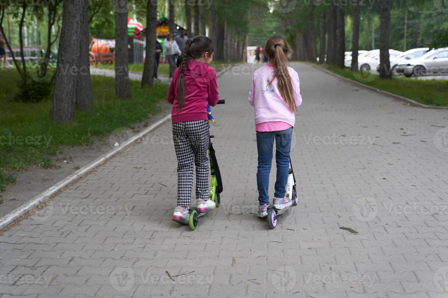 View from behind of unrecognizable girls riding kick scooters on alley, pavement on summer day, with copy space. Concept of friendship, sport, healthy and active lifestyle, eco transport, fun photo