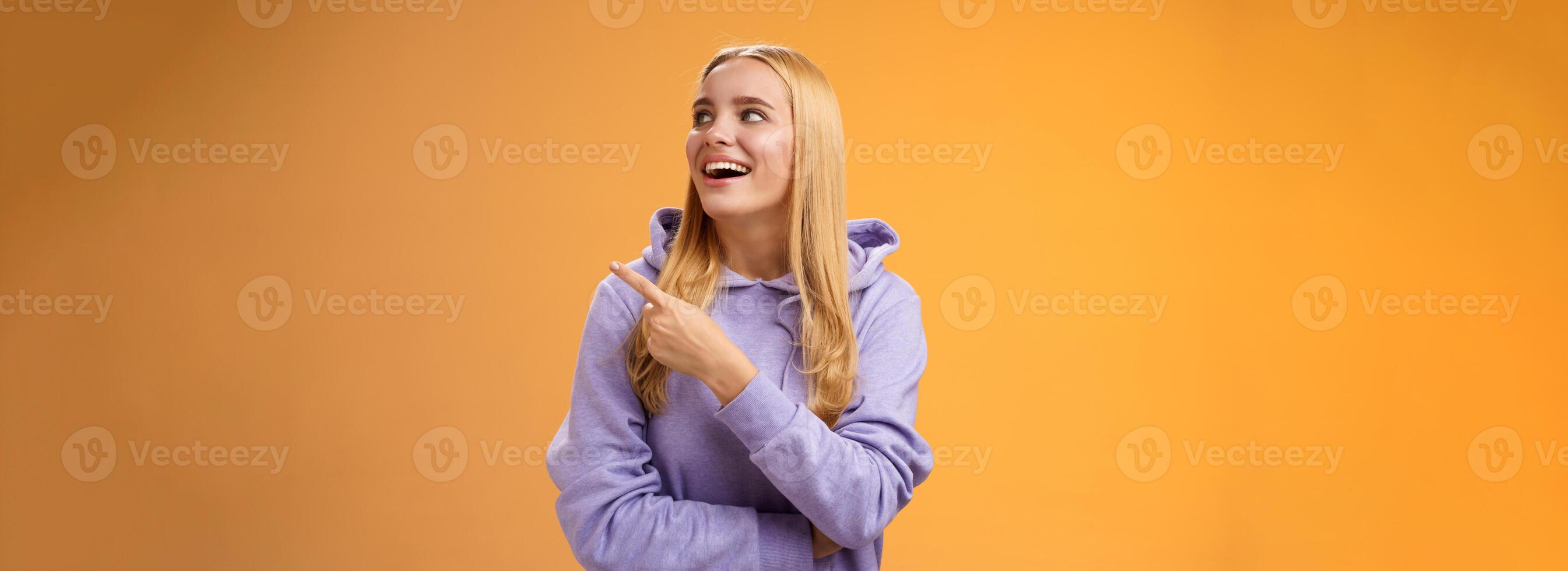 Amused carefree joyful blond european woman in hoodie pointing looking right pleased smiling happily enjoying interesting fascinating peromance making choice in store shopping, orange background photo