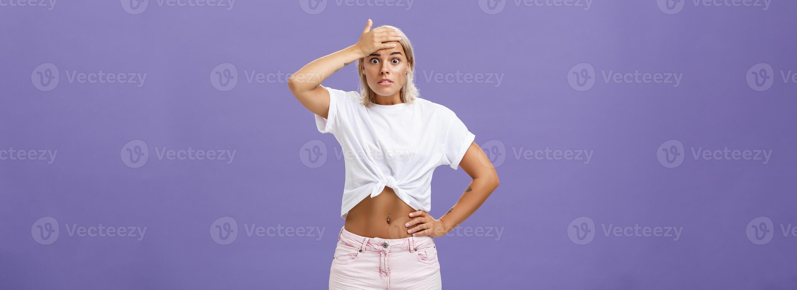 Damn I am in trouble. Worried troubled good-looking stylish female employee with tanned skin and fit body holding hand on forehead popping eyes from anxiety and making nervous face over purple wall photo
