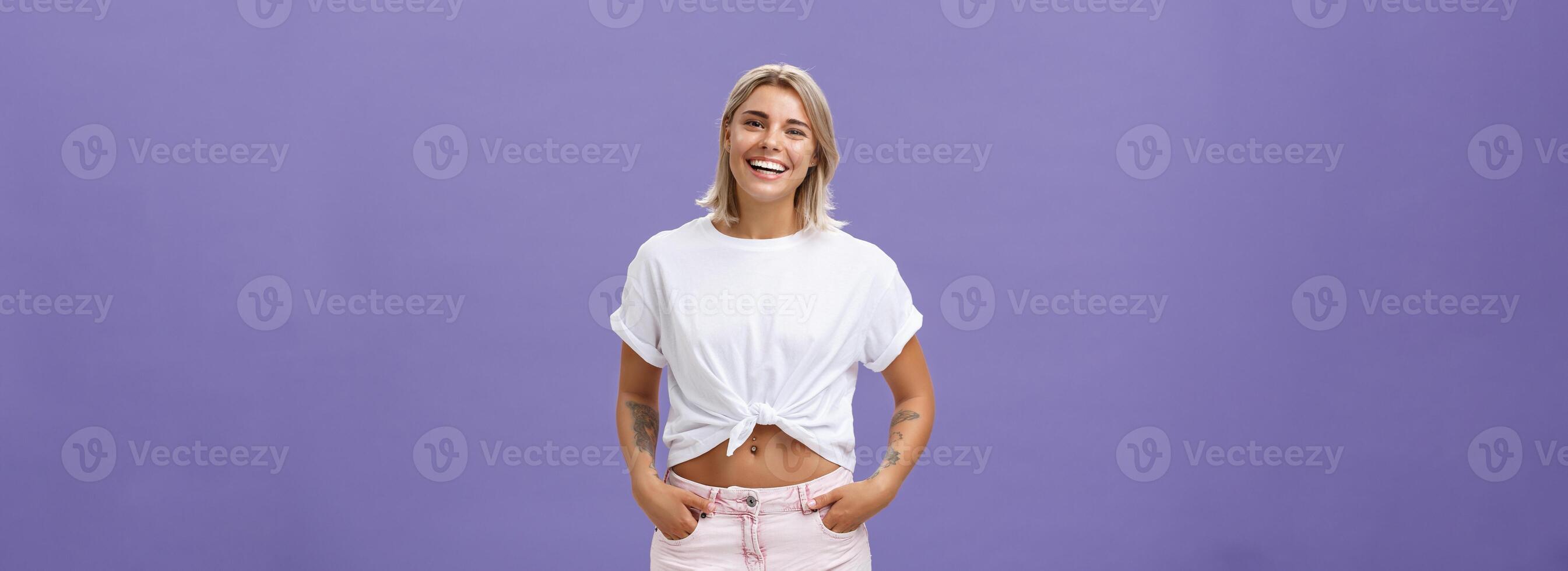 Joyful attractive tanned girlfriend with blond hair and tattoos on arms with cropped top chilling by pool with friends talking with amused happy expression smiling at camera holding hands in pockets photo