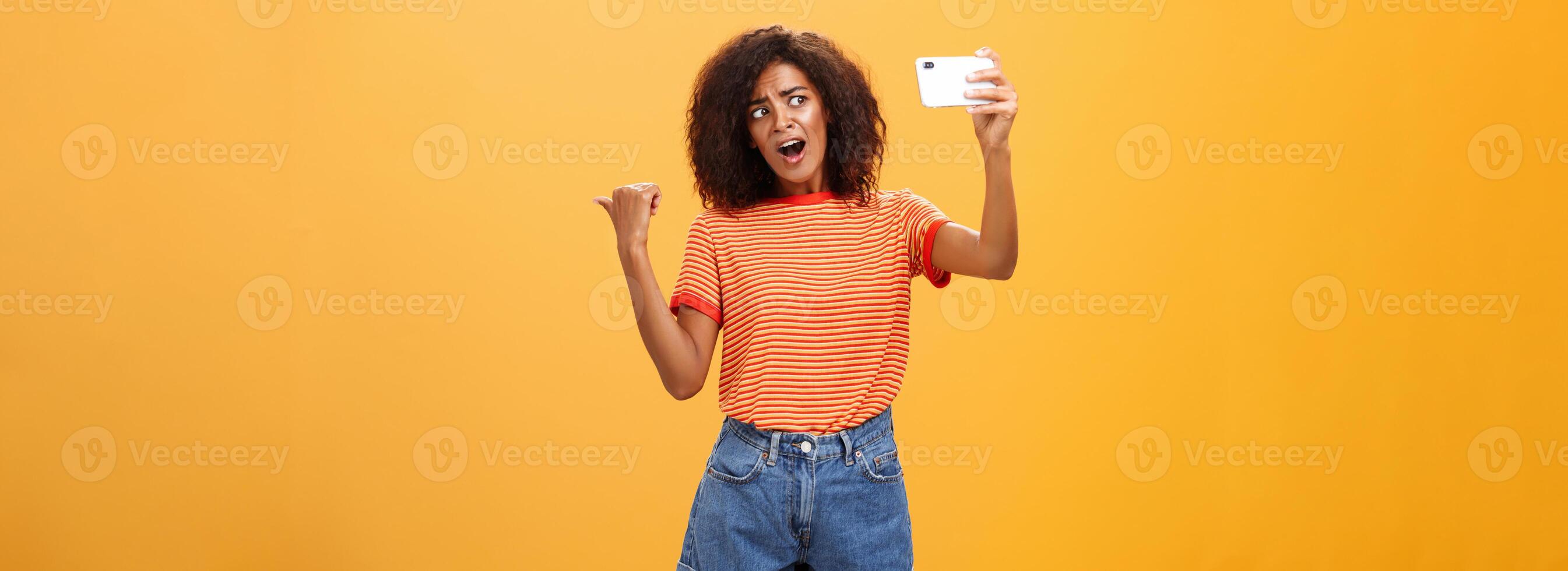 Woman recording blog pointing at strange object behind her. Portrait of concerned and curious stylish famous internet star holding smartphone talking in cellphone camera over orange background photo