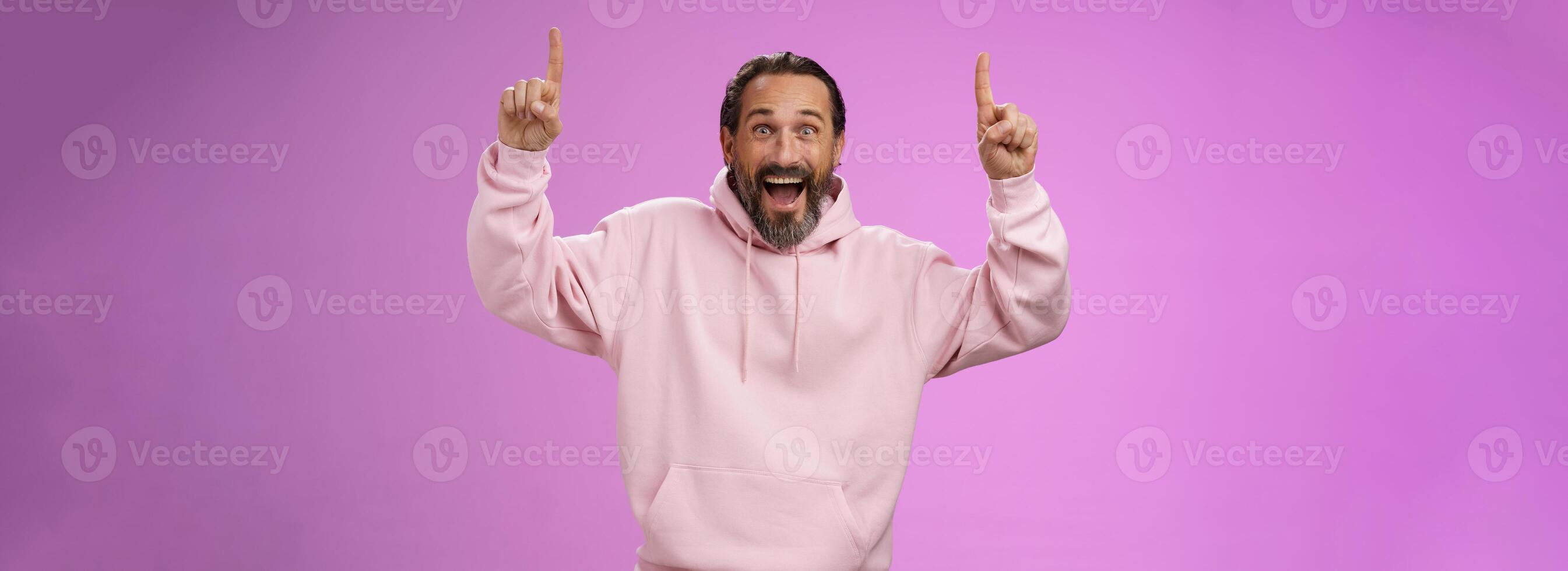 Energized happy good-looking trendy mature dad beard grey hair in pink stylish hoodie wearing son outfit pointing up index fingers thrilled favorite music group in town, standing purple background photo