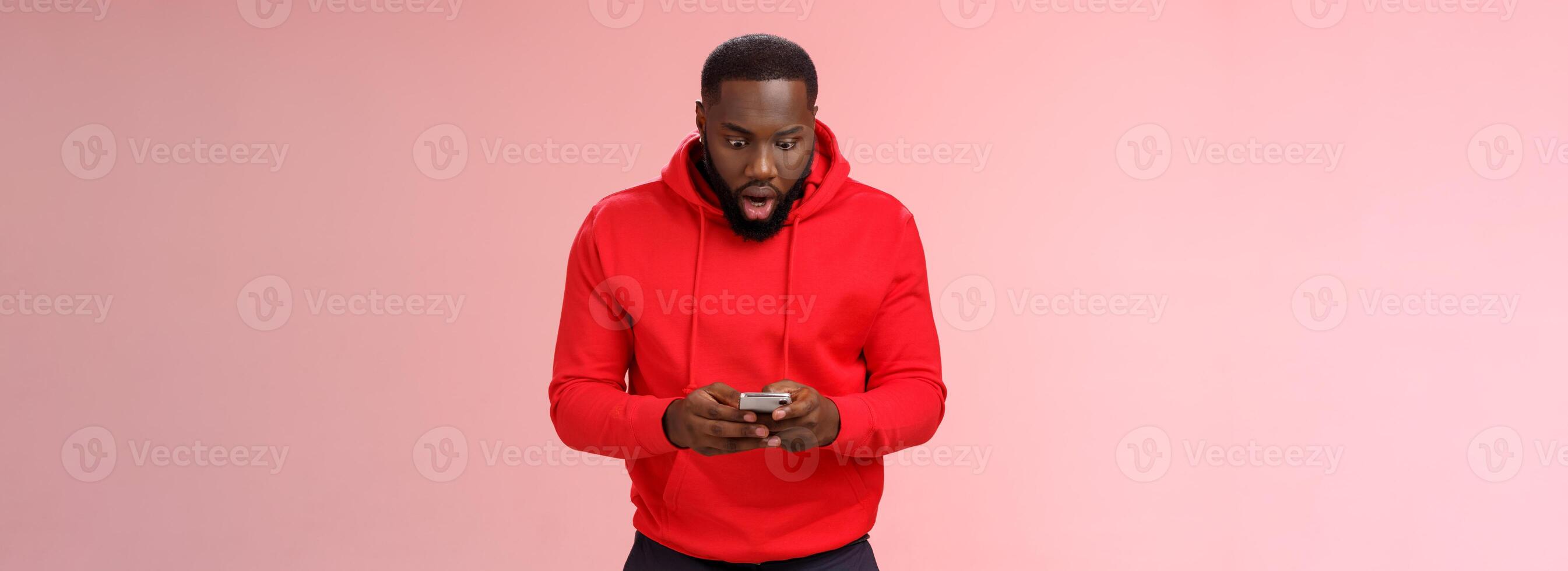 Amused focused impressed african-american bearded guy in red hoodie drop jaw widen eyes look amazed smartphone display winning hard game level answering exciting message, standing pink background photo